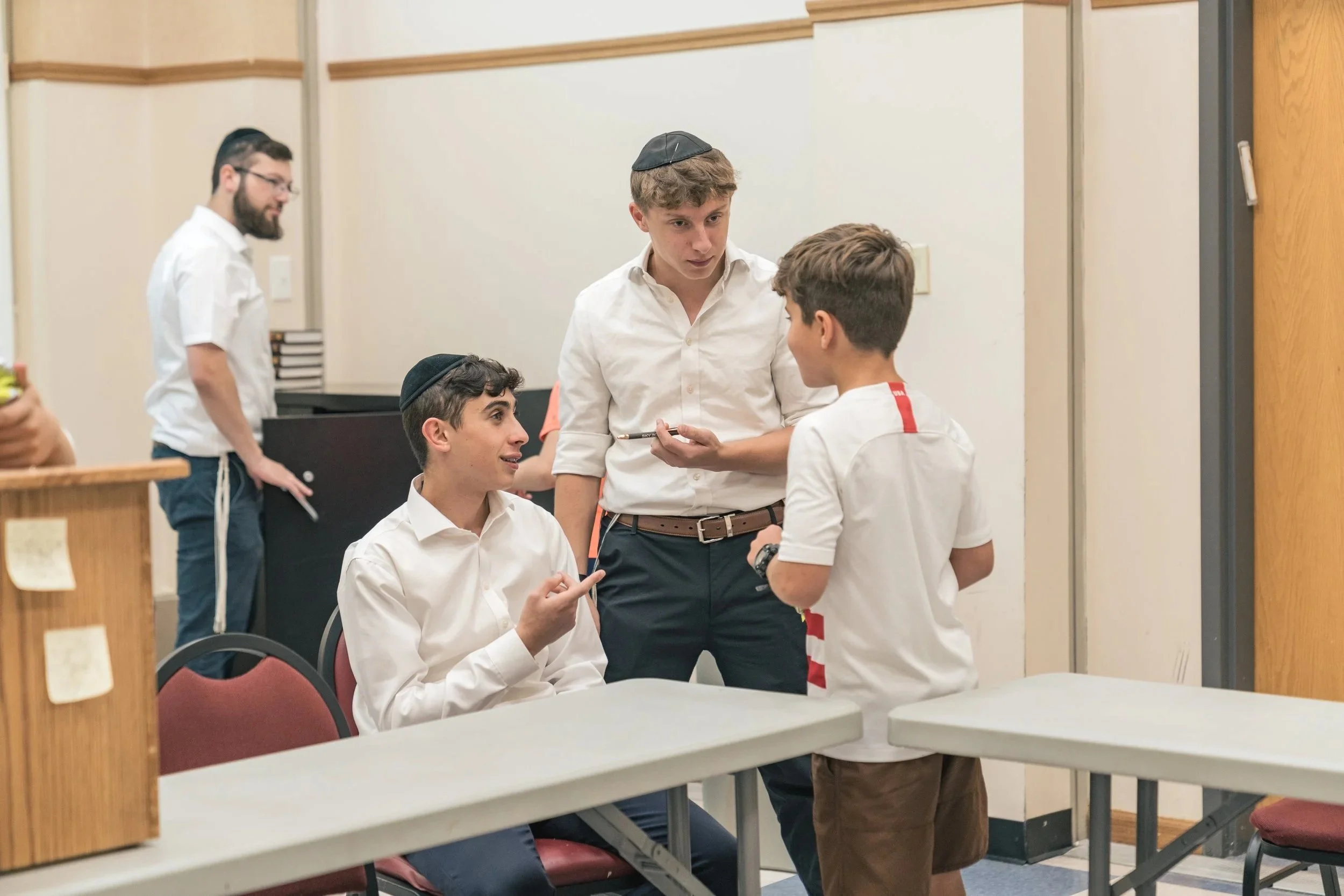 Four young boys in white shirts and yarmulkes engaging in conversation in a classroom or meeting room.