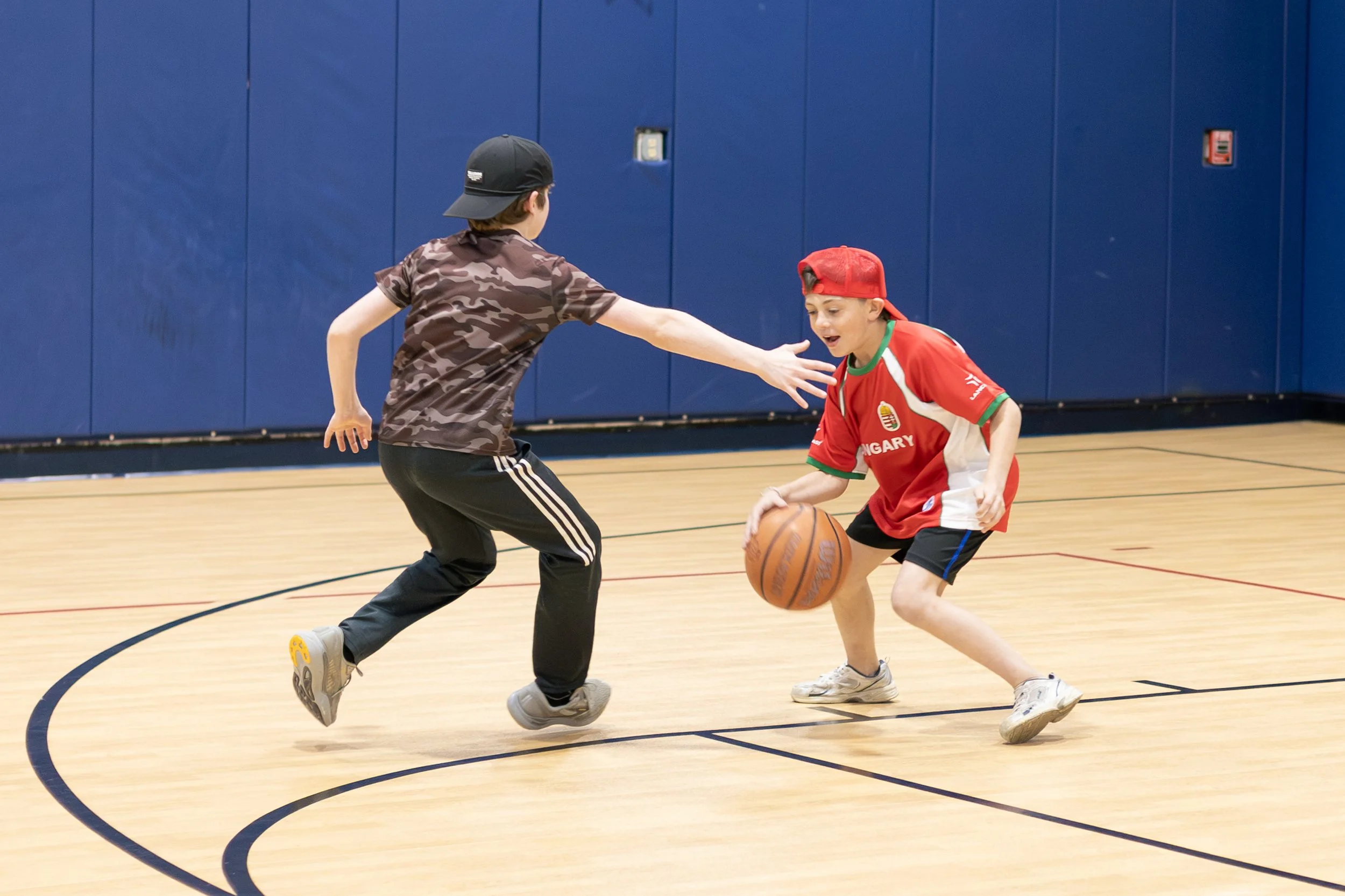 Two boys playing basketball in an indoor gym, one in a camouflage shirt and black cap reaching for the ball, the other in a red sports jersey and cap attempting to dribble.