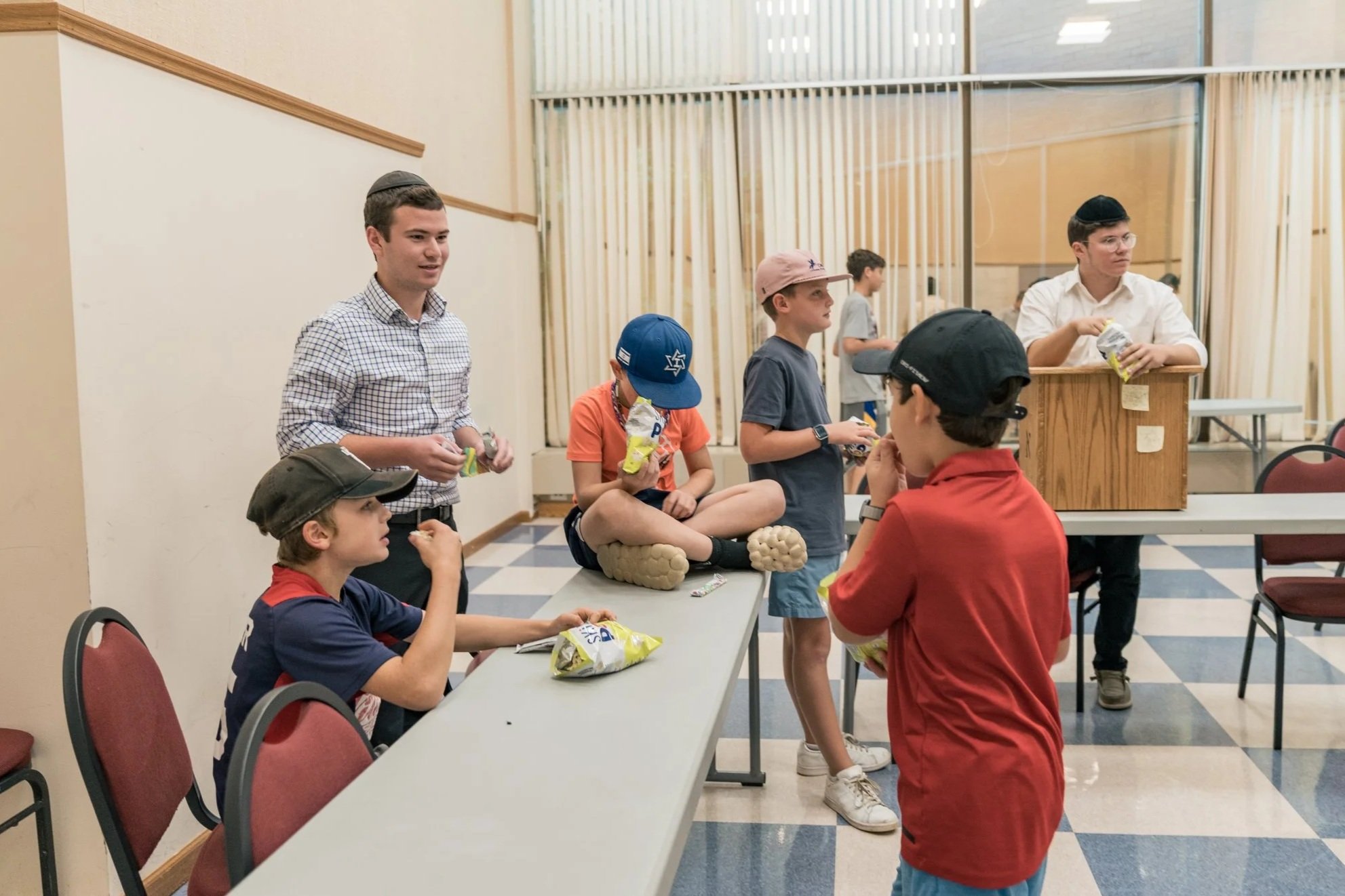 Young boys and men in a room with tables and chairs, some wearing kippahs, gathering snacks, with one sitting cross-legged on the table, and others eating or preparing snacks.