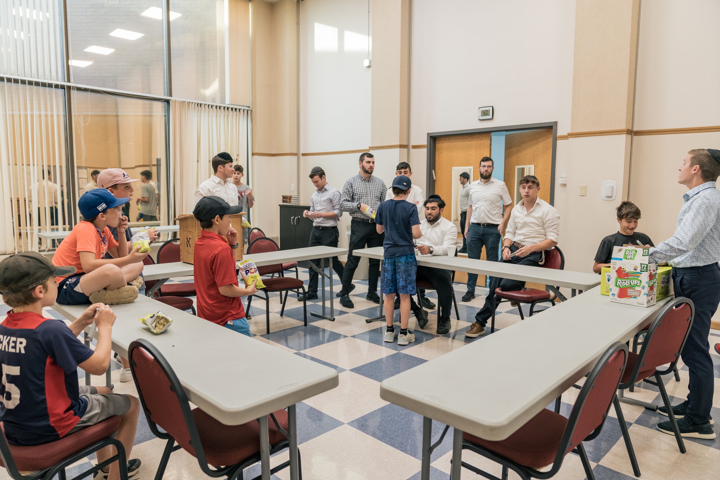 Children and adults gathered around tables in a room, engaging in an activity, with snacks on the tables and some adults standing and sitting.
