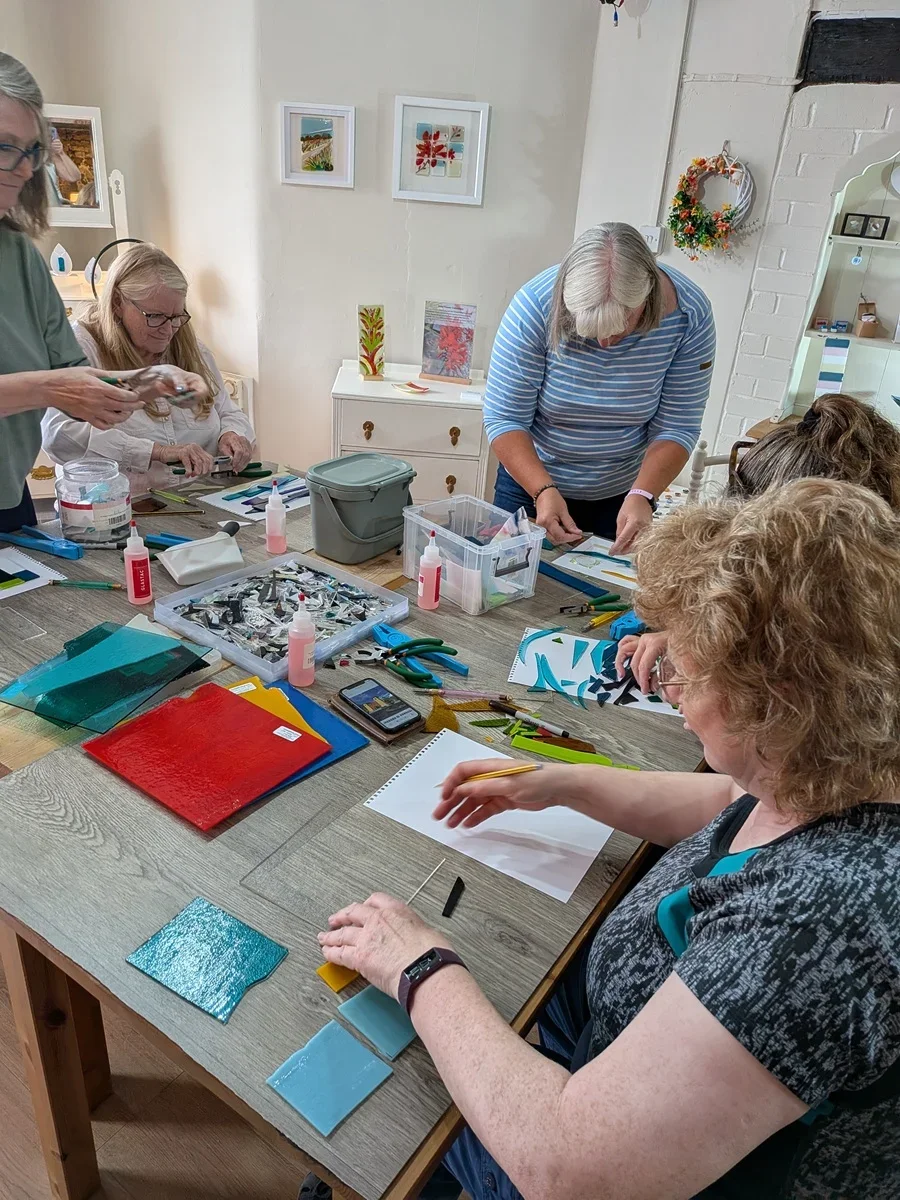 A group of women working on glass art projects at a workshop table, with various glass pieces, tools, and supplies laid out.