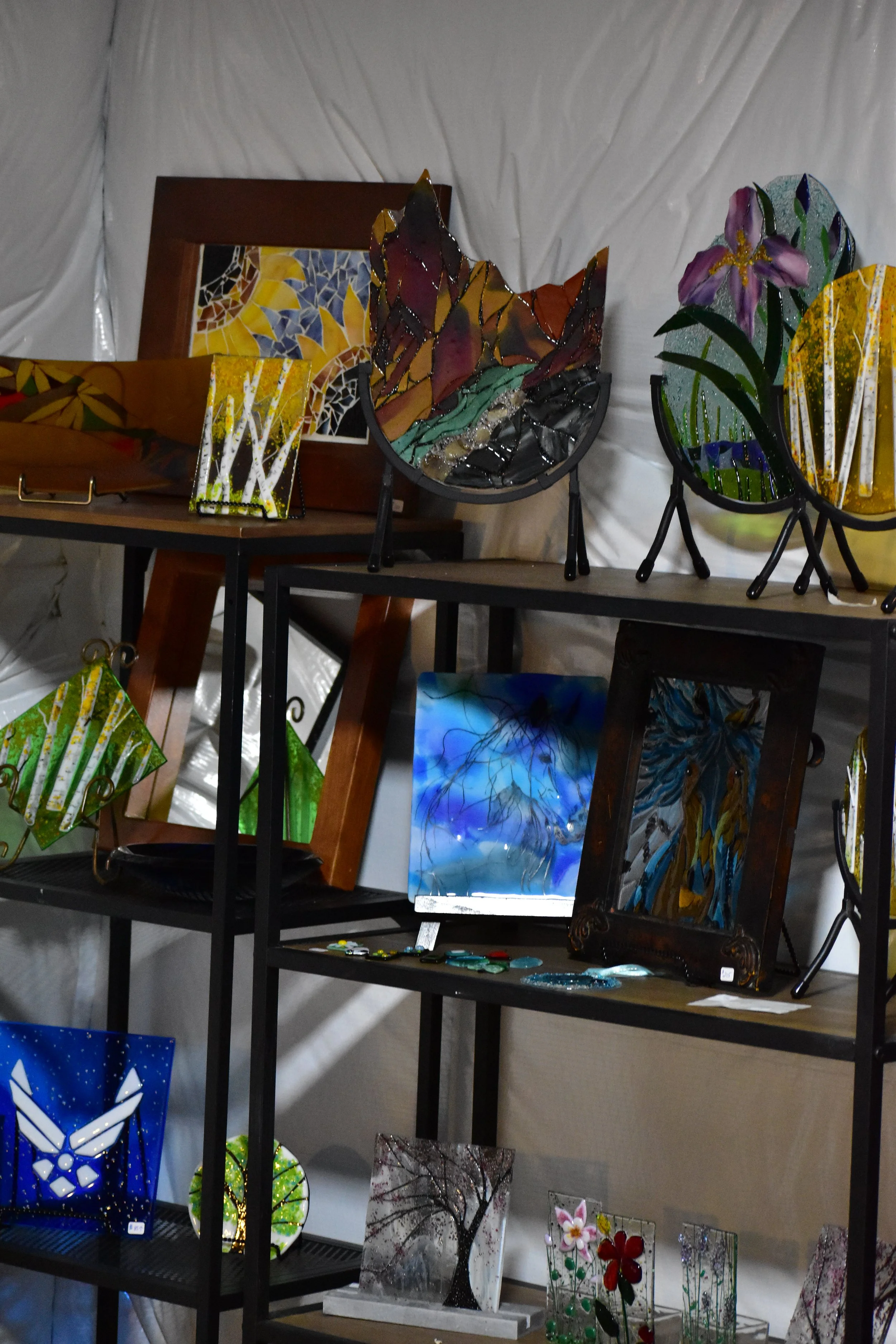 Display of colorful glass art pieces including stained glass tiles, framed pictures, and decorative sculptures on shelves and stands at an art fair or gallery.