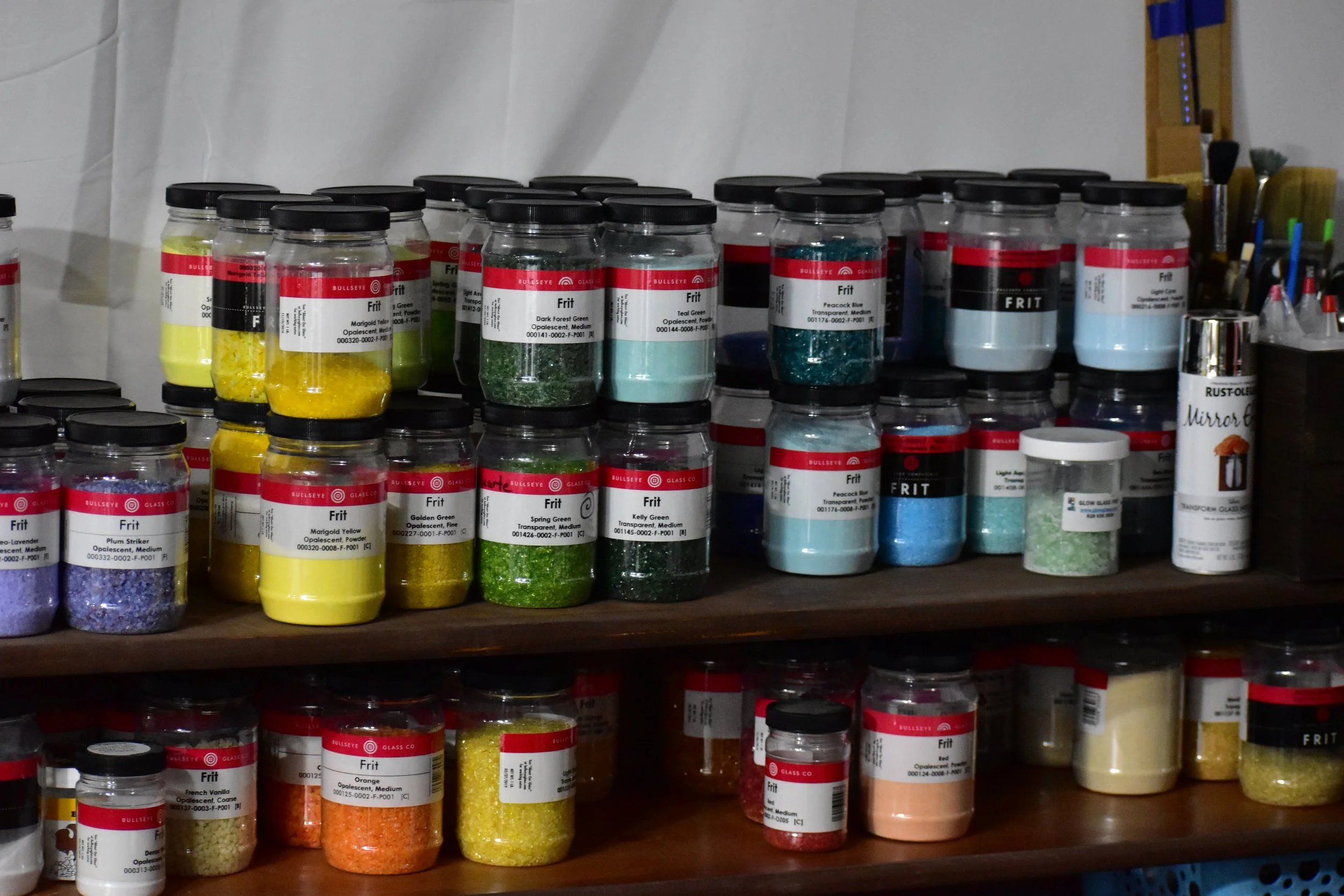 Shelves filled with jars of colored glitters and powder used for crafting, along with a container of green glitter and other small supplies.