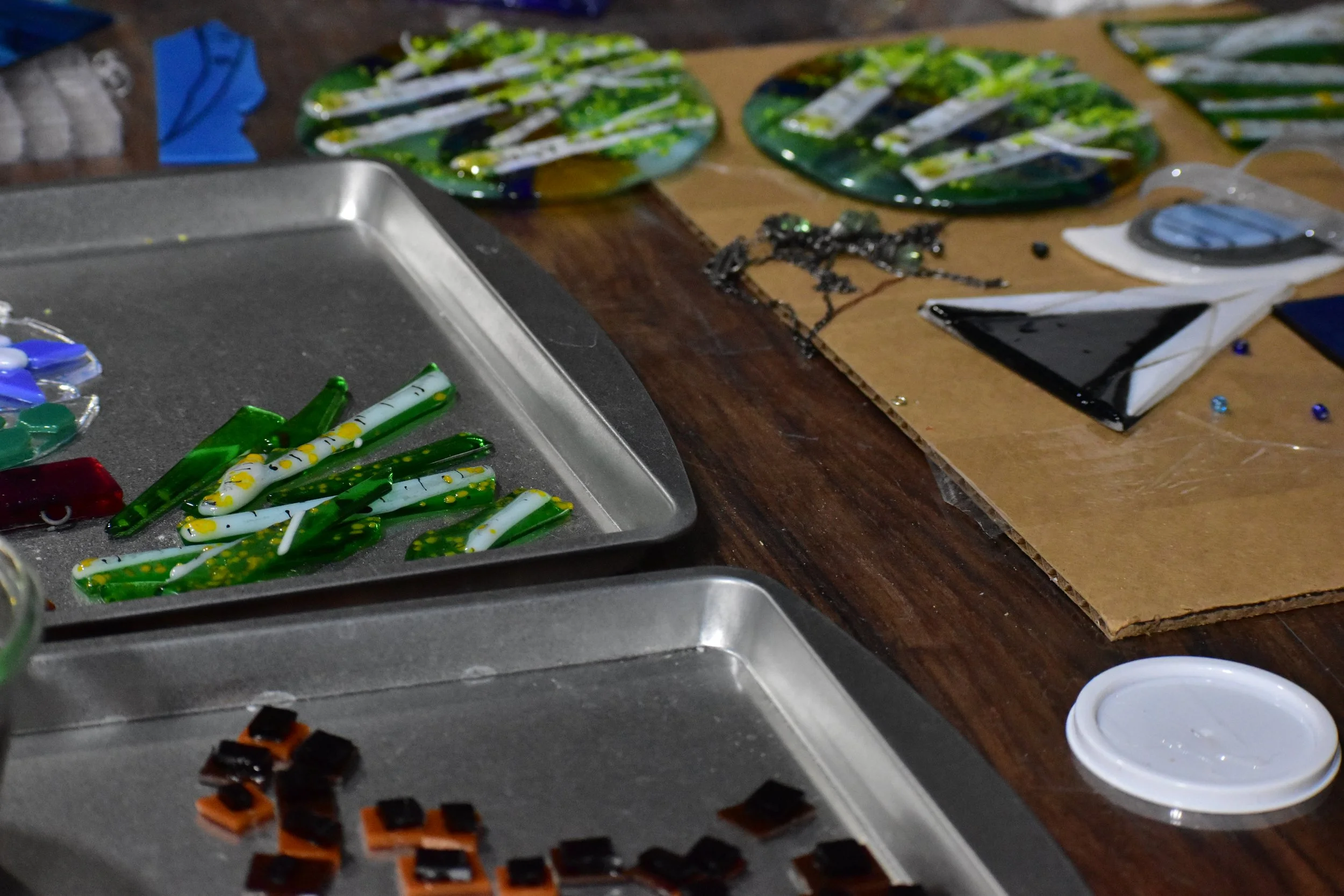 A workspace with glass mosaic pieces in various colors and shapes, some on metal trays and others on paper, used for creating stained glass or mosaic art.