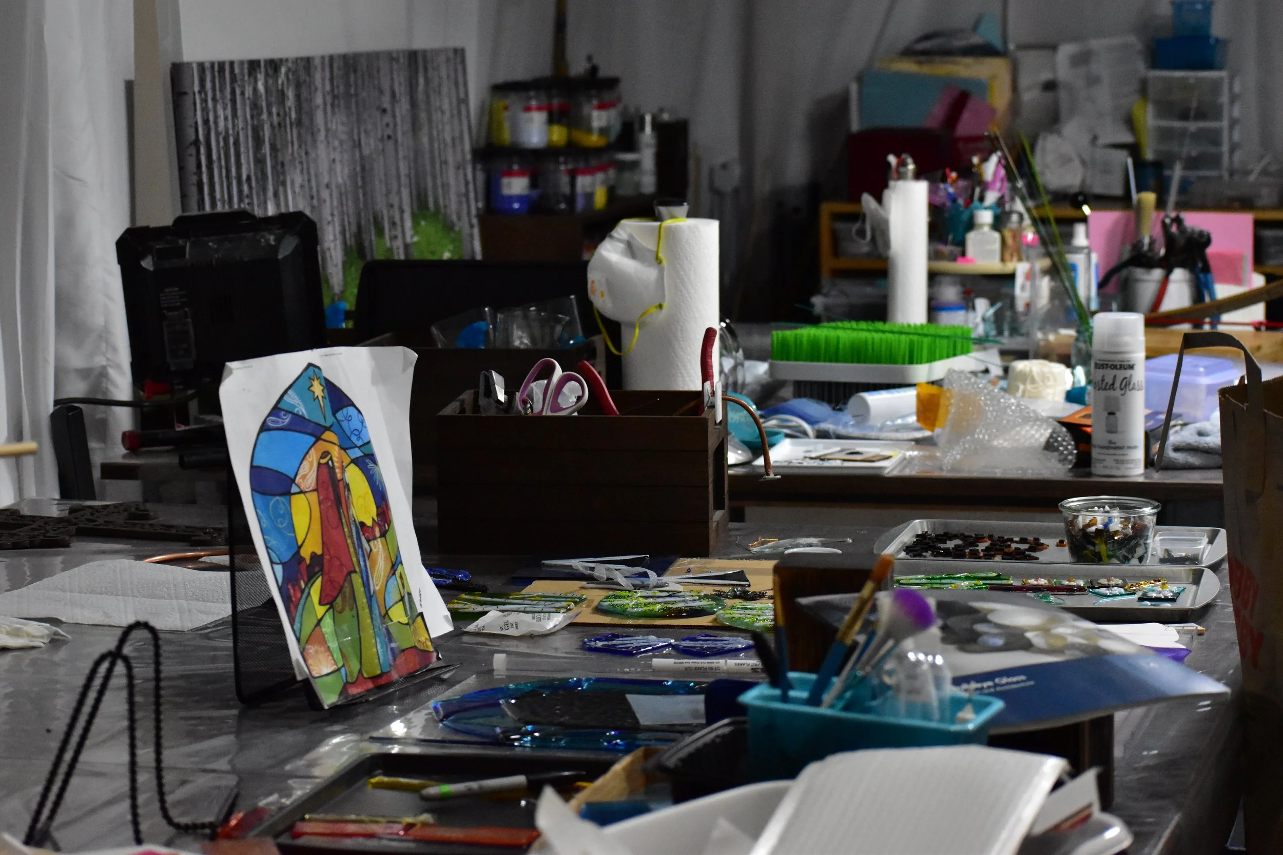 A cluttered workspace with various craft supplies, including paints, scissors, paper, and stained glass pieces, on tables and shelves.