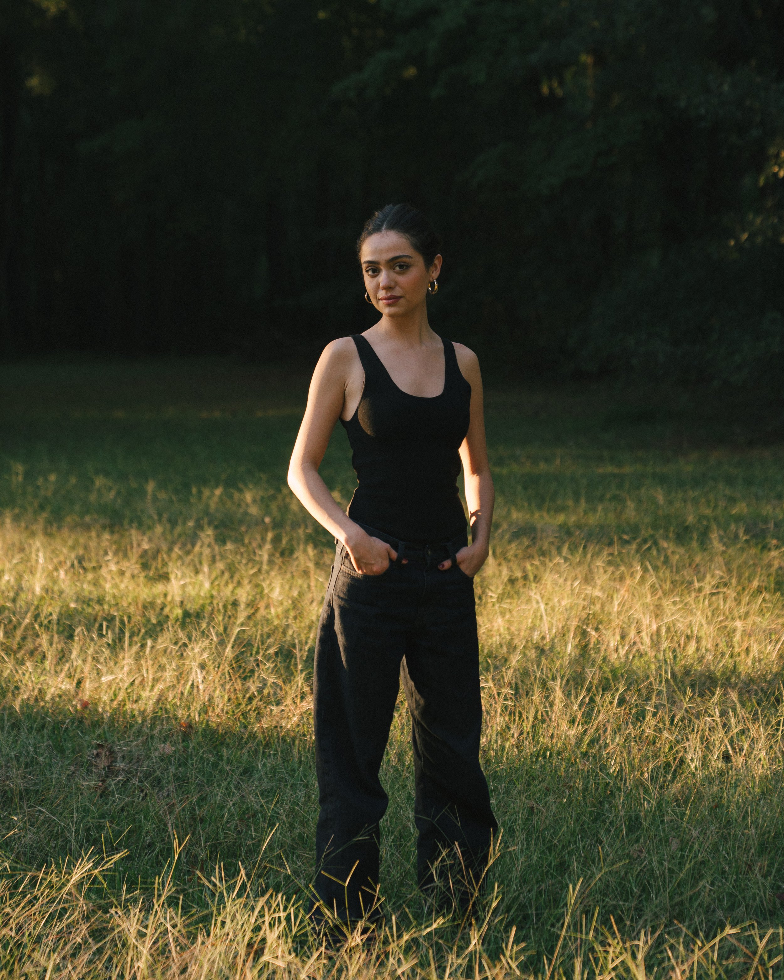 A young woman with dark hair tied back, wearing a sleeveless black top and black pants, stands in a grassy field during sunset with her hands in her pockets, looking at the camera.