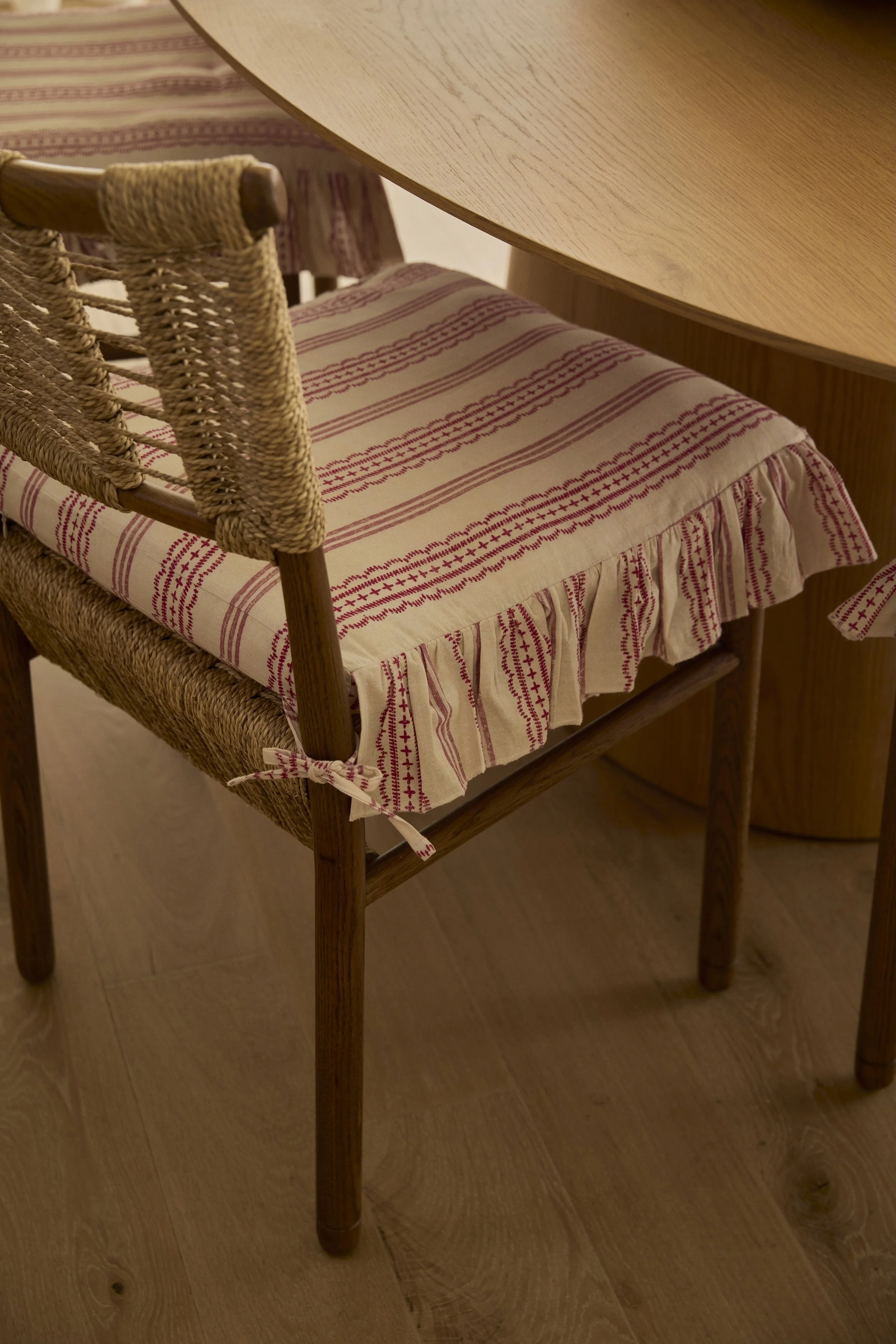 Wooden dining chair with woven seat and a fabric cushion with red stripes and ruffles, next to a wooden table on a light wood floor.