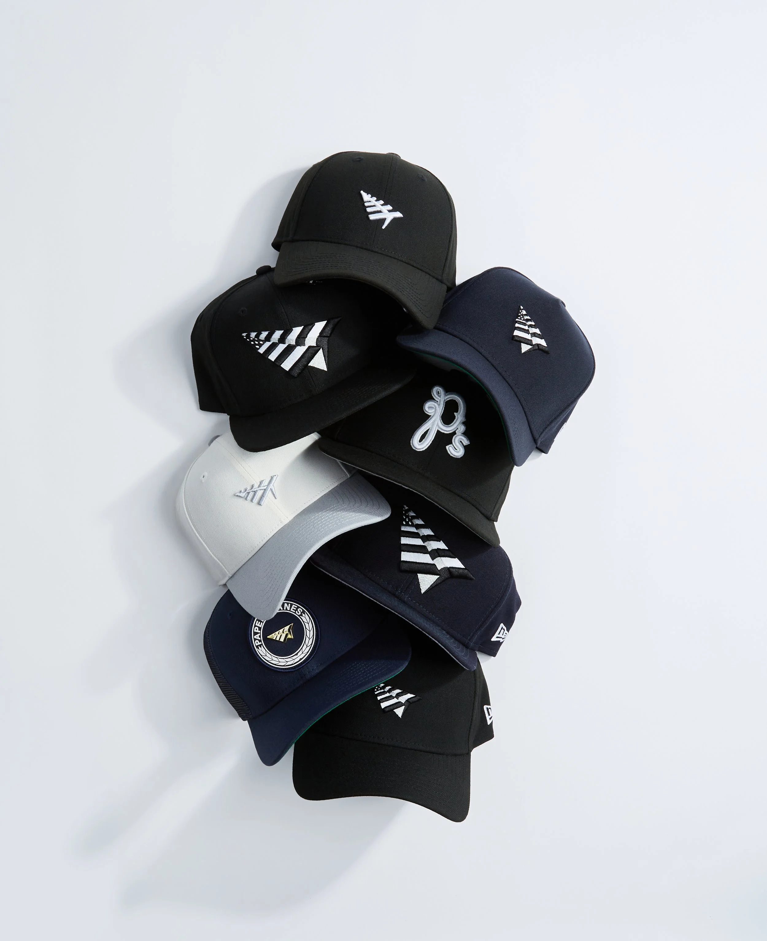 A collection of six baseball caps in black, white, navy blue, and dark gray, each with a white striped logo, laid out on a white surface.