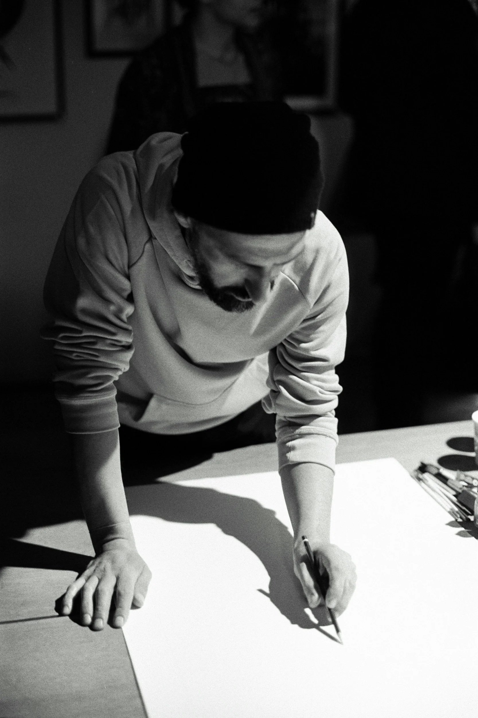 Black-and-white photo of a man with a beard and mustache, wearing a beanie and a light-colored sweatshirt, drawing or writing on a large piece of paper on a table, with art supplies and pens nearby.