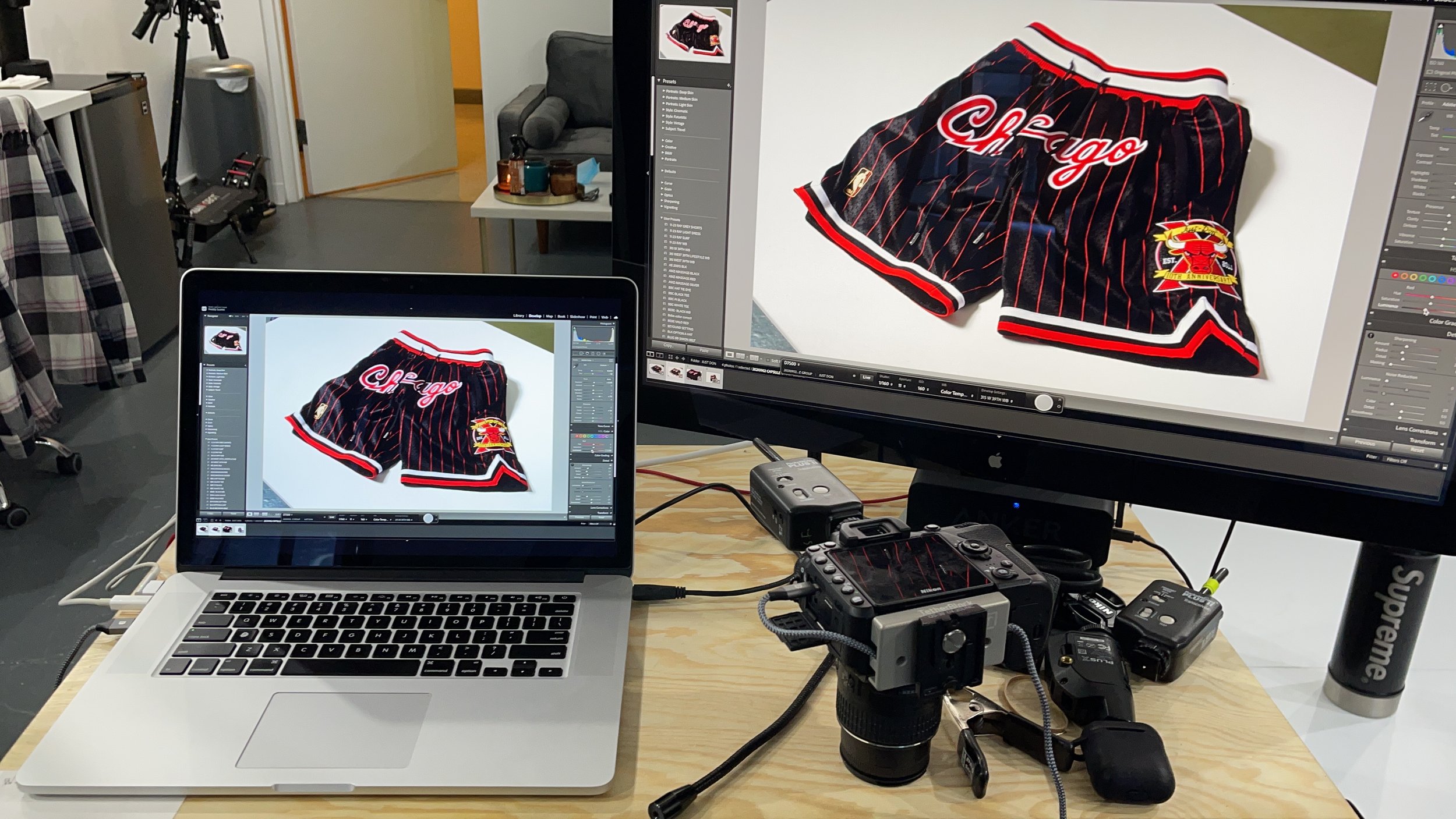 A workspace with a MacBook laptop and a second monitor displaying a photo editing software. The photo shows a pair of Chicago Bulls basketball shorts with red and black pinstripes, a Bulls logo, and the word 'Chicago' on the front. Various photography equipment and cables are on the table, and in the background, there is a gray chair, a tripod, and a small table with candles and objects.