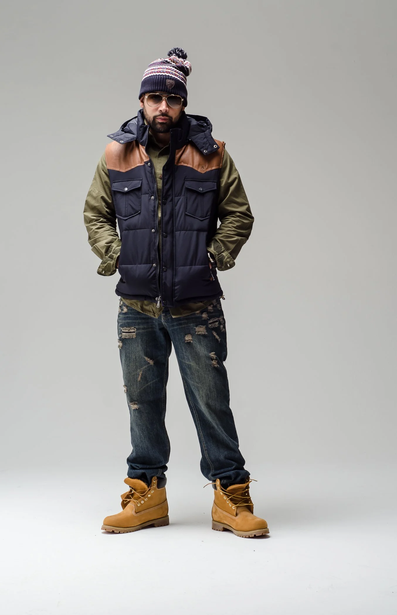 A man standing with hands in his pockets, dressed in casual winter clothing, wearing a knit hat, sunglasses, a layered vest over a shirt, distressed jeans, and tan work boots, against a plain background.
