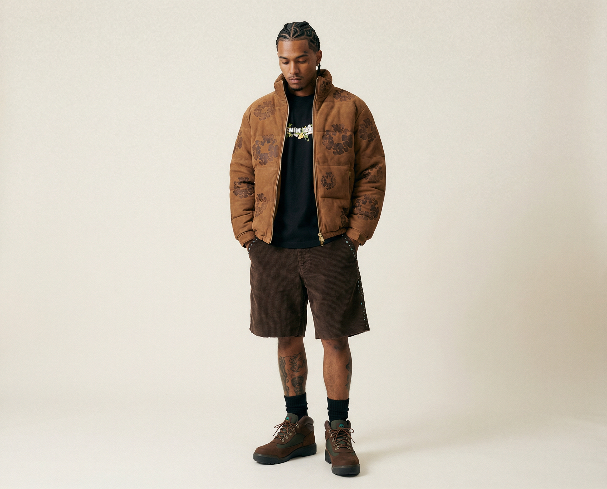 A young man with braided hairstyle wearing a brown patterned jacket, black T-shirt, brown shorts, and hiking boots standing against a plain light background.