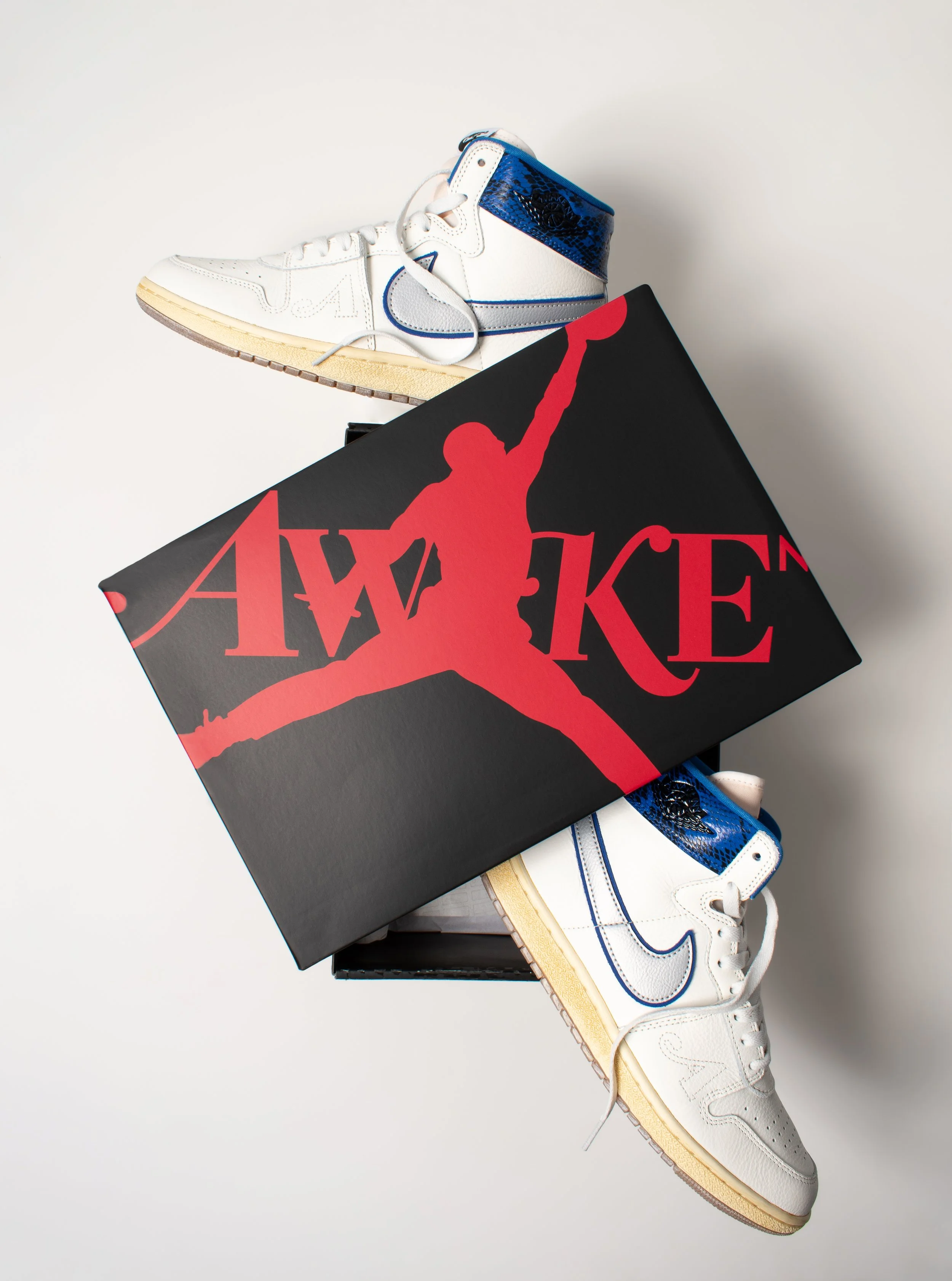 A white Nike high-top sneaker with blue accents and a blue snakeskin pattern on the ankle area, placed on top of a black box with a red logo of a man jumping with a basketball, set against a white background.