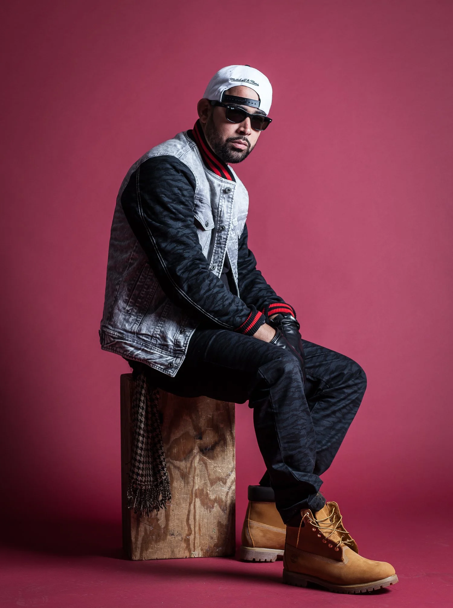A man sitting on a wooden block against a pink background, wearing sunglasses, a white baseball cap backwards, a denim jacket with black patterned sleeves, dark pants, and tan work boots.