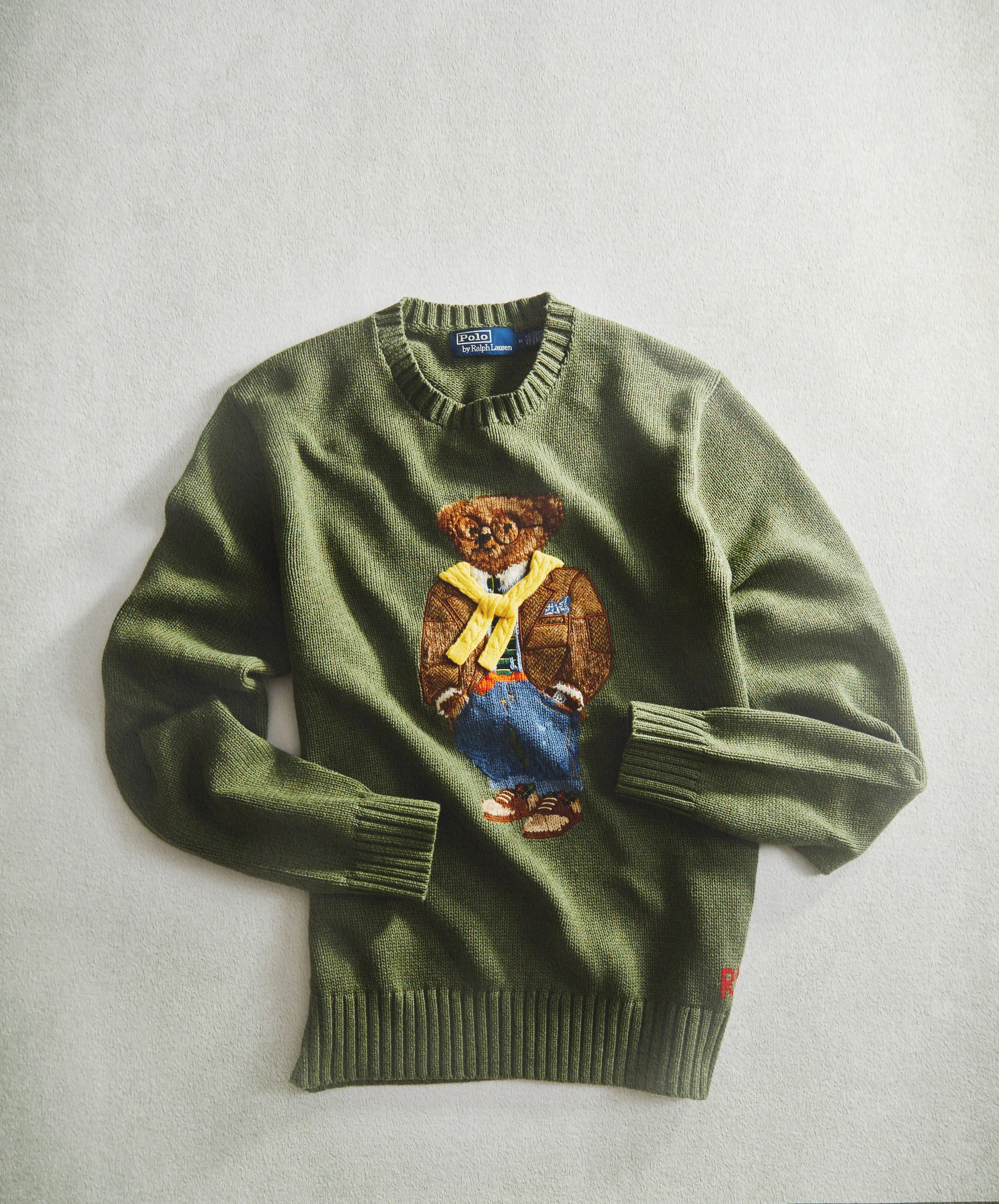 Green knitted sweater featuring an embroidered teddy bear wearing a yellow shirt and blue jeans, with a red 'R' visible on the bottom right.