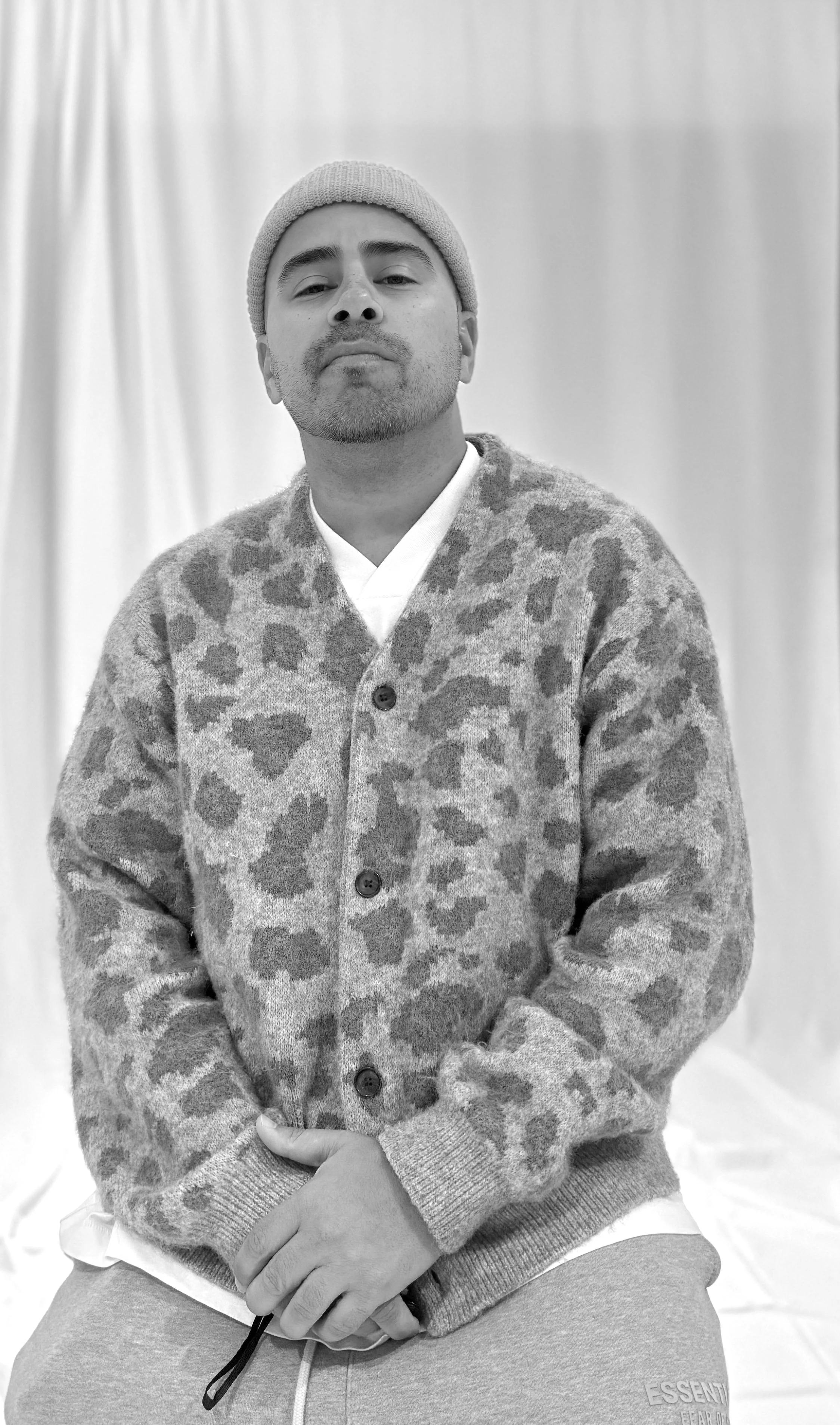Black and white photo of a man wearing a knit beanie, a patterned cardigan, a white shirt, and sweatpants, sitting against a plain background.
