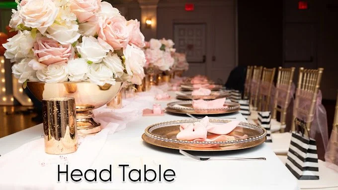 Elegant head table decorated with large floral arrangements in pink and white, set with pink and gold plates, silverware, and pink napkins, with gold chairs and black-and-white striped party favor bags.