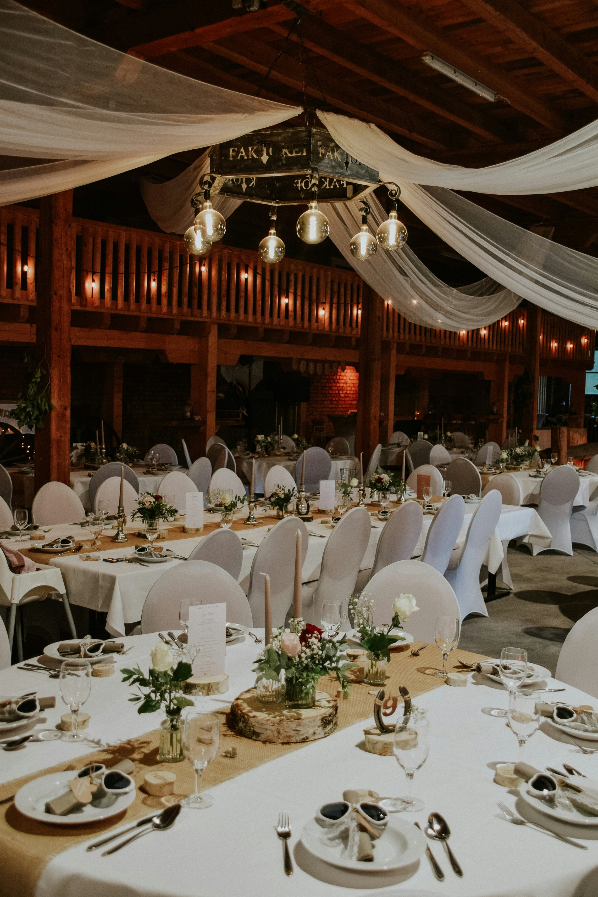 Indoor banquet hall decorated with white drapes and string lights, featuring long tables with white tablecloths, floral centerpieces, and place settings for a formal event.