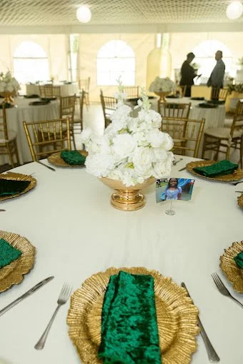 Elegant event table setup with a white floral centerpiece, gold chargers, green napkins, and gold flatware, in a bright venue with large windows and two people conversing in the background.
