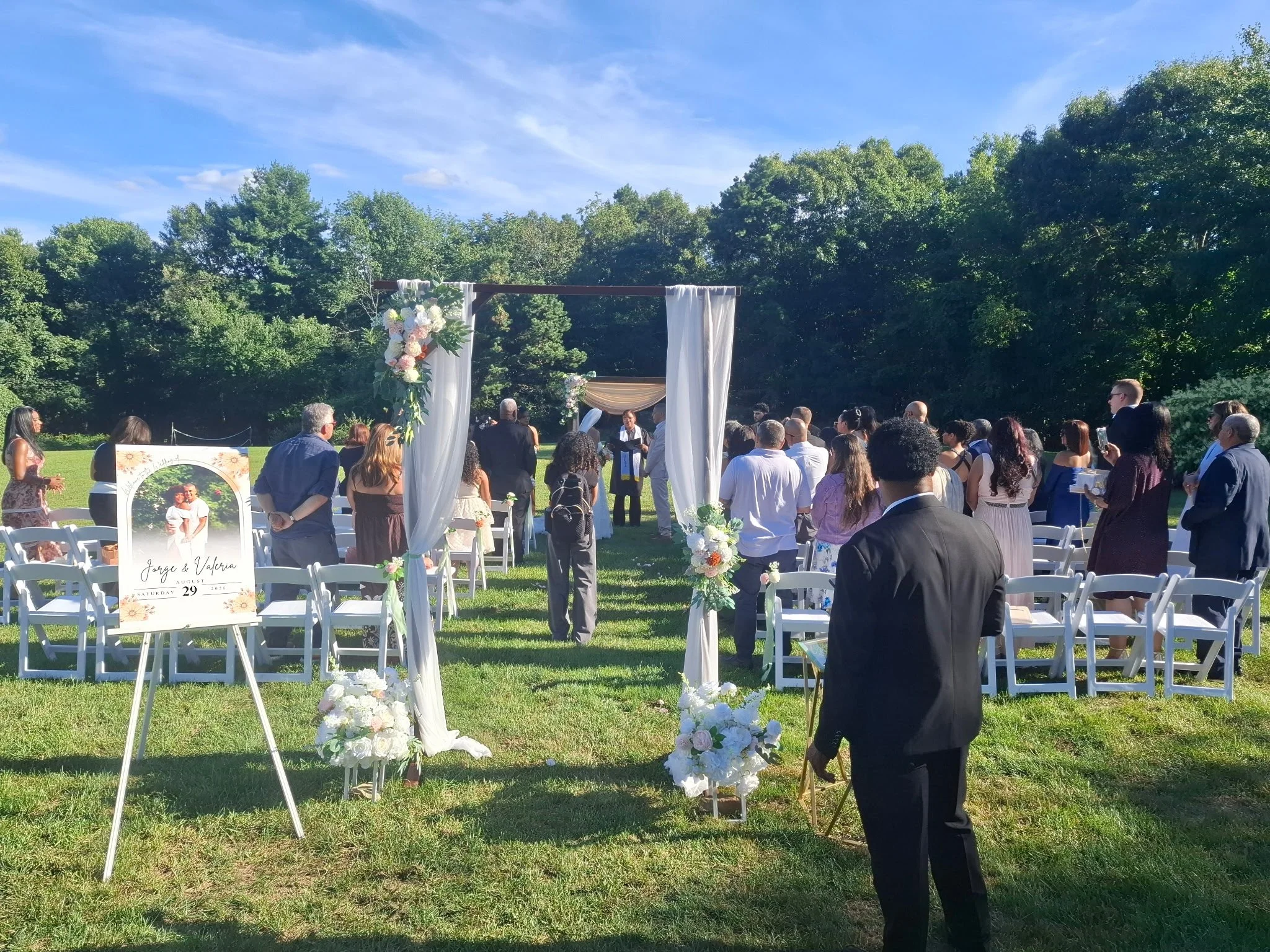 An outdoor wedding ceremony setup with white chairs arranged on a grassy field, decorated with white flowers, in front of an archway adorned with flowers and draped white fabric. Guests are gathered, and the sky is blue with some clouds.
