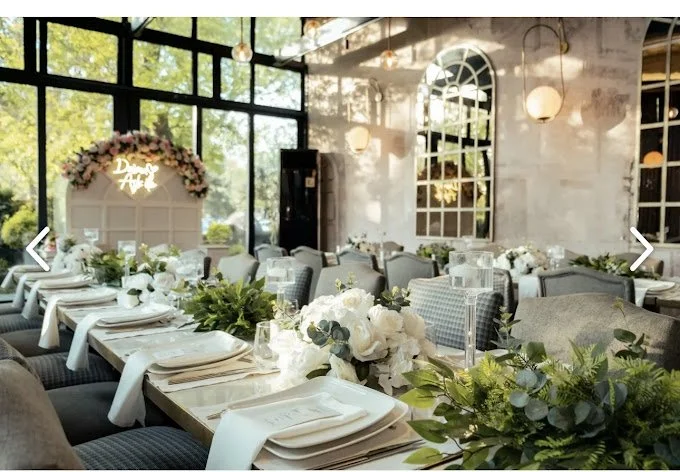 Wedding reception decorated with white flowers and greenery, set with white napkins and plates, in a bright room with large windows and hanging pendant lights.