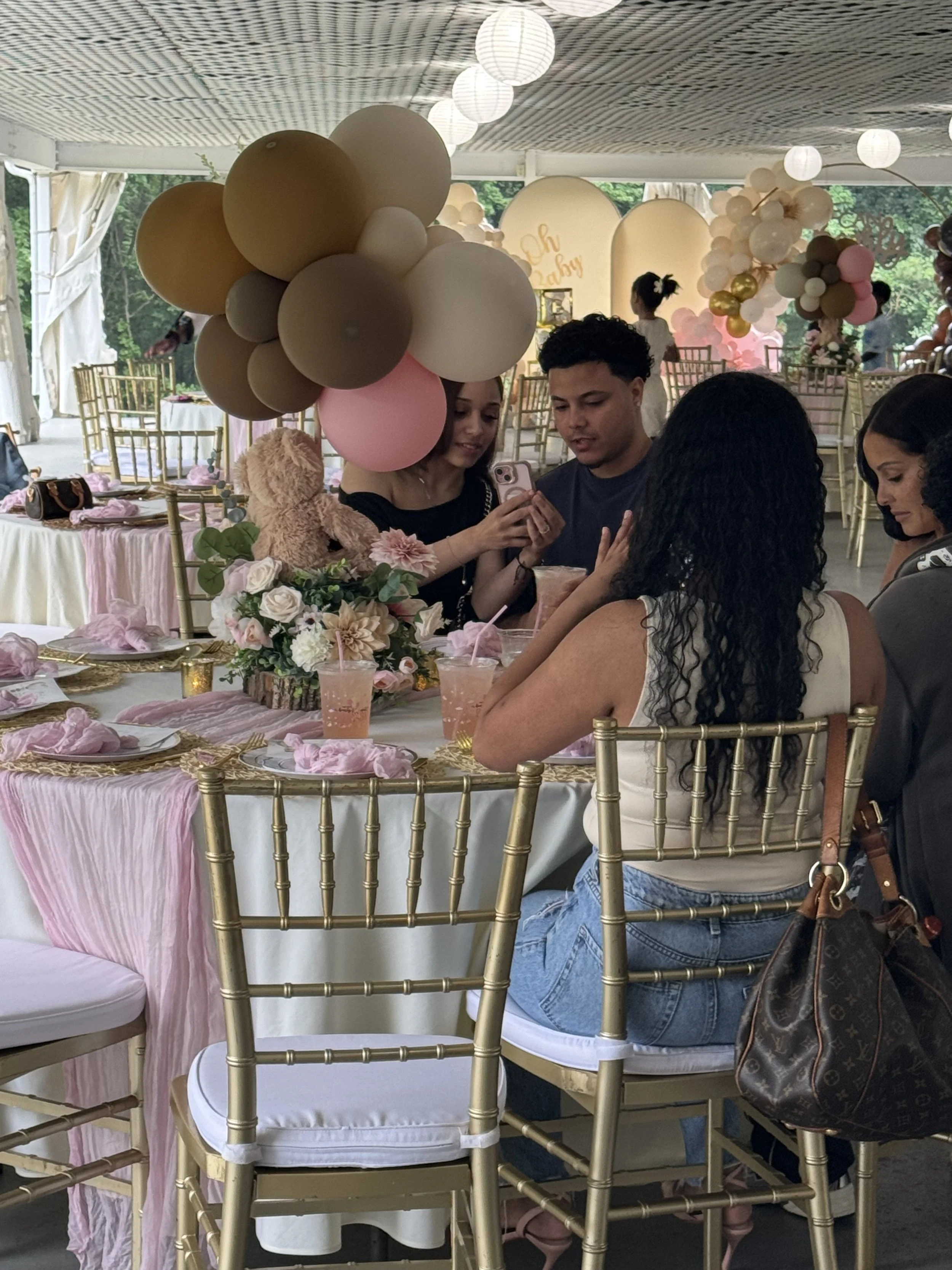 People sitting at a decorated table in a party or celebration setting, with balloons, floral arrangements, and pink accents.