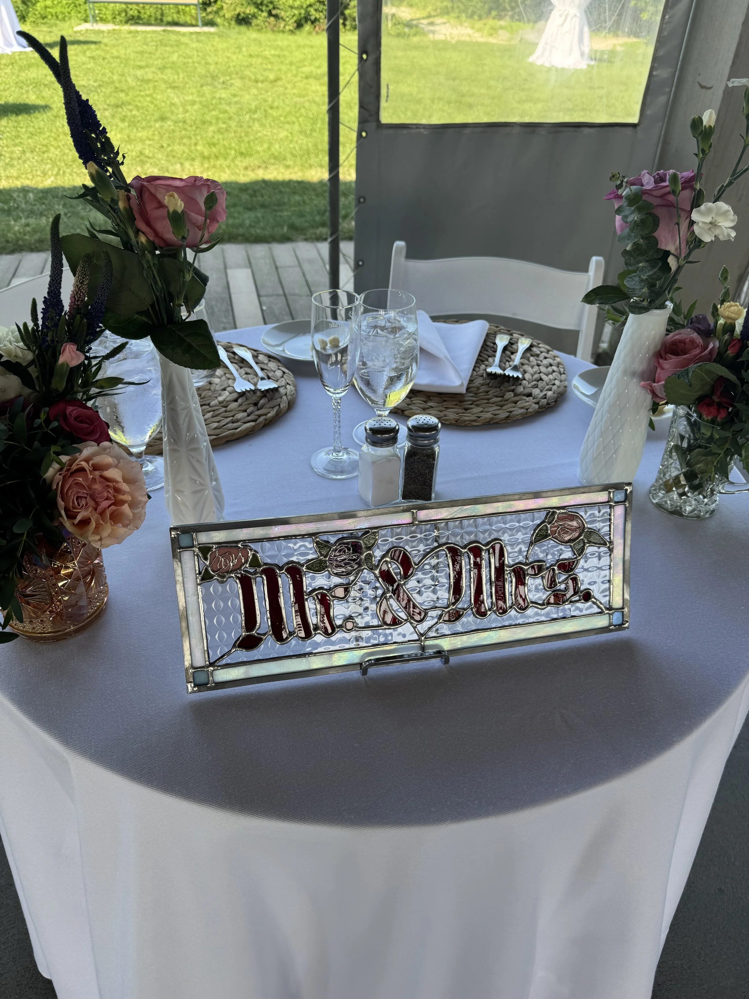 A table decorated with pink and purple flowers in vases, set for a meal with plates, cutlery, like forks and knives, glasses of water and champagne, surrounded by a white tablecloth and a decorative metal sign that says 'Mr. & Mrs.' in front of an ou