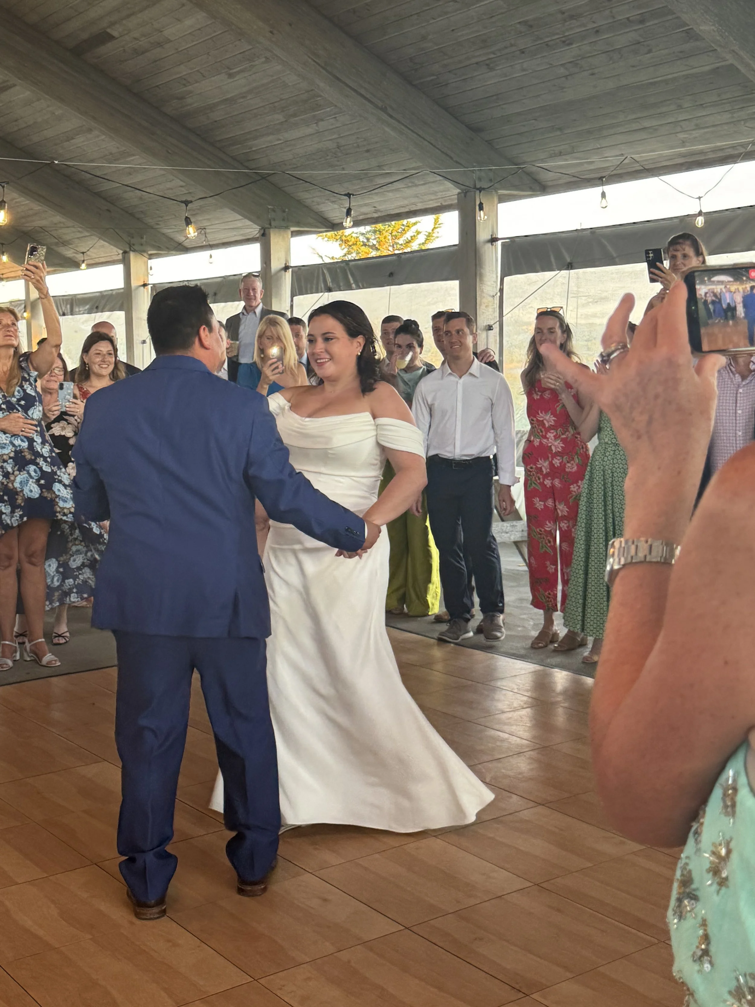 A bride and groom dancing at their wedding reception with guests surrounding them, some taking photos.