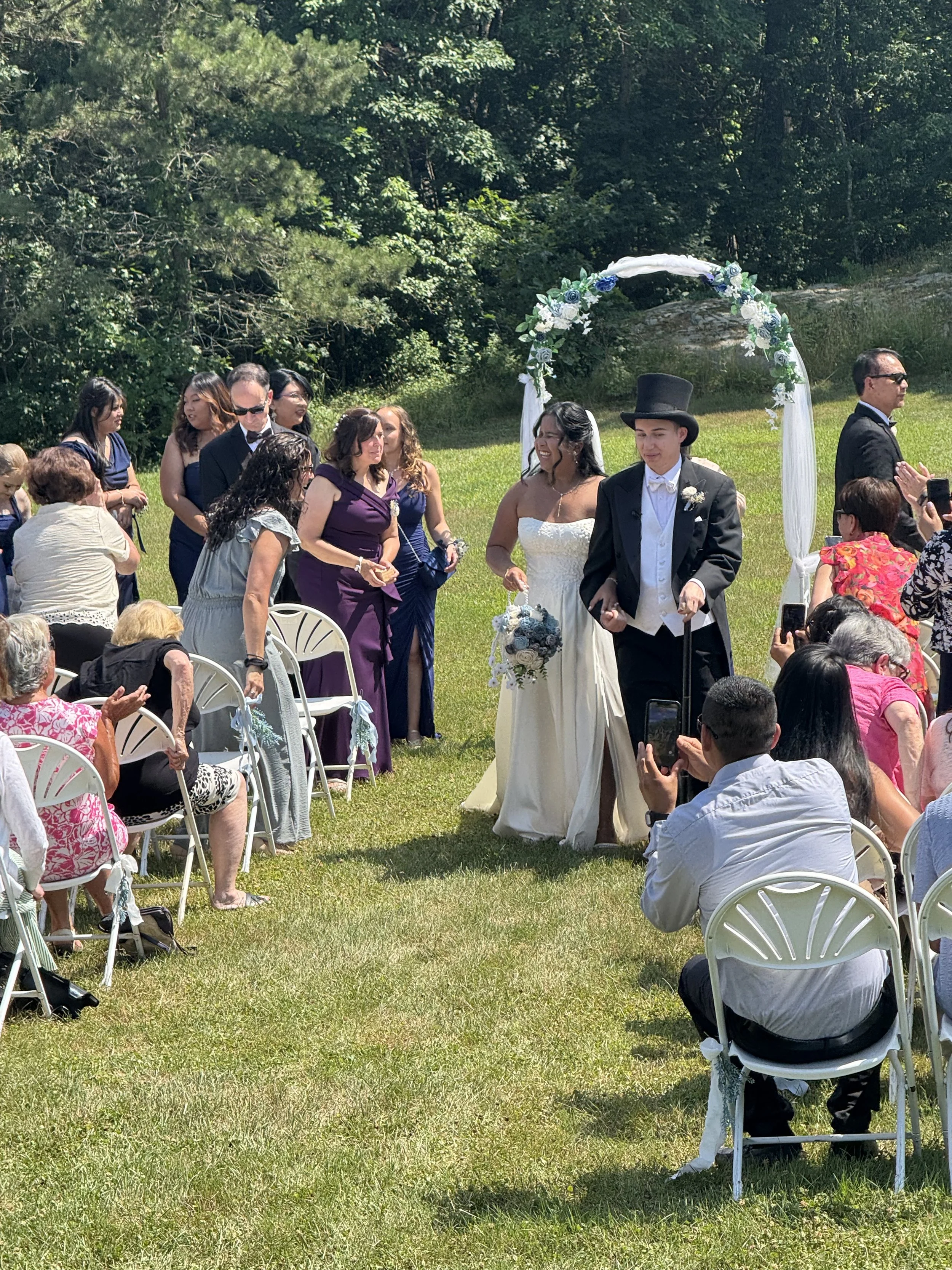 Outdoor wedding ceremony with a bride and groom walking under a floral arch, surrounded by seated guests on a grassy area with trees in the background.