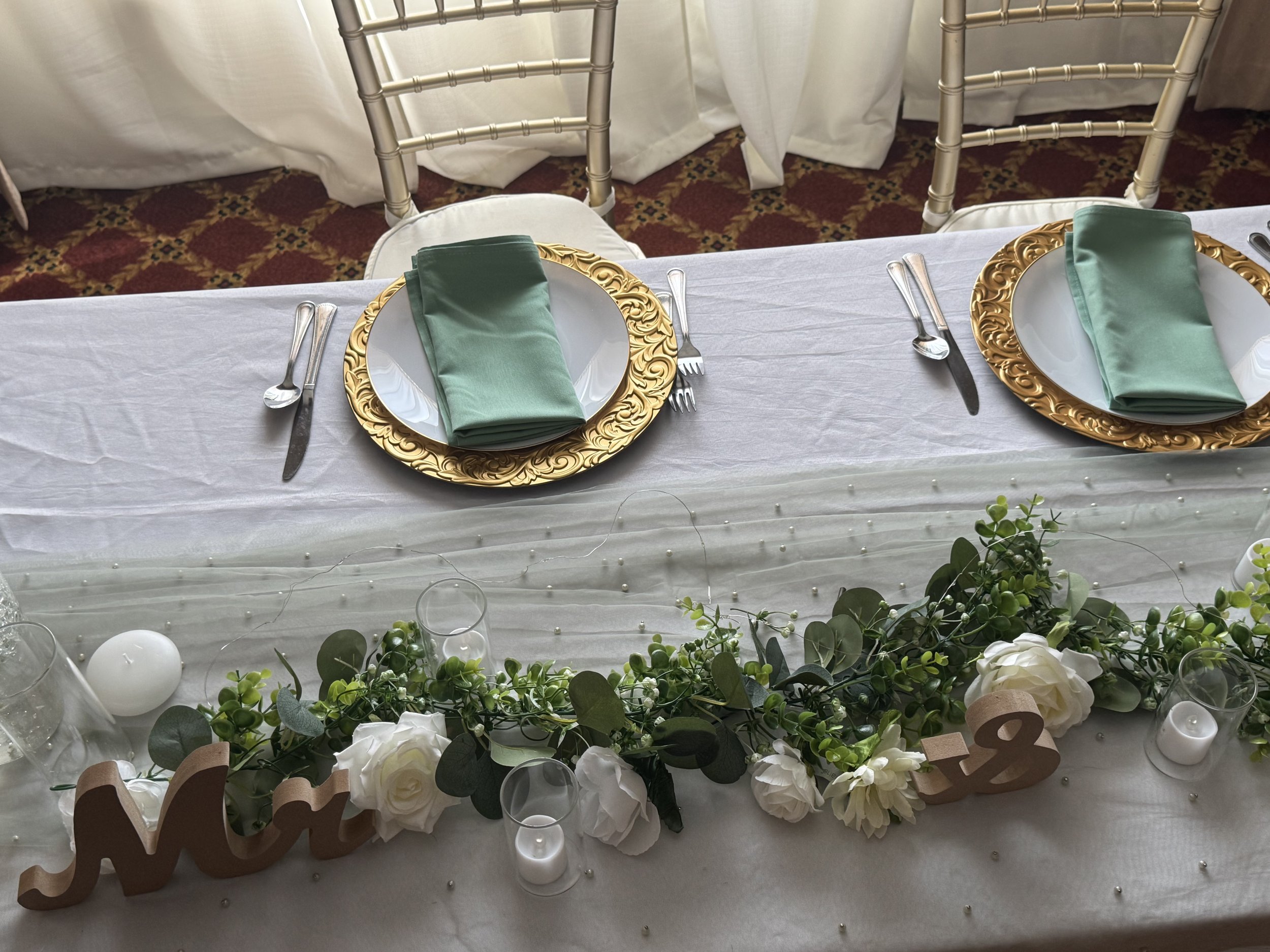 A table set for a formal event with two place settings, each with a white plate, gold decorative charger, green napkin, fork, knife, and spoon. The table is decorated with white flowers, green foliage, candles, and a number '02' sign, with chairs in 