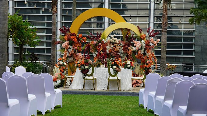 Outdoor wedding ceremony setup with floral arch, chairs covered in white, and decorative yellow rings in front of a modern building.