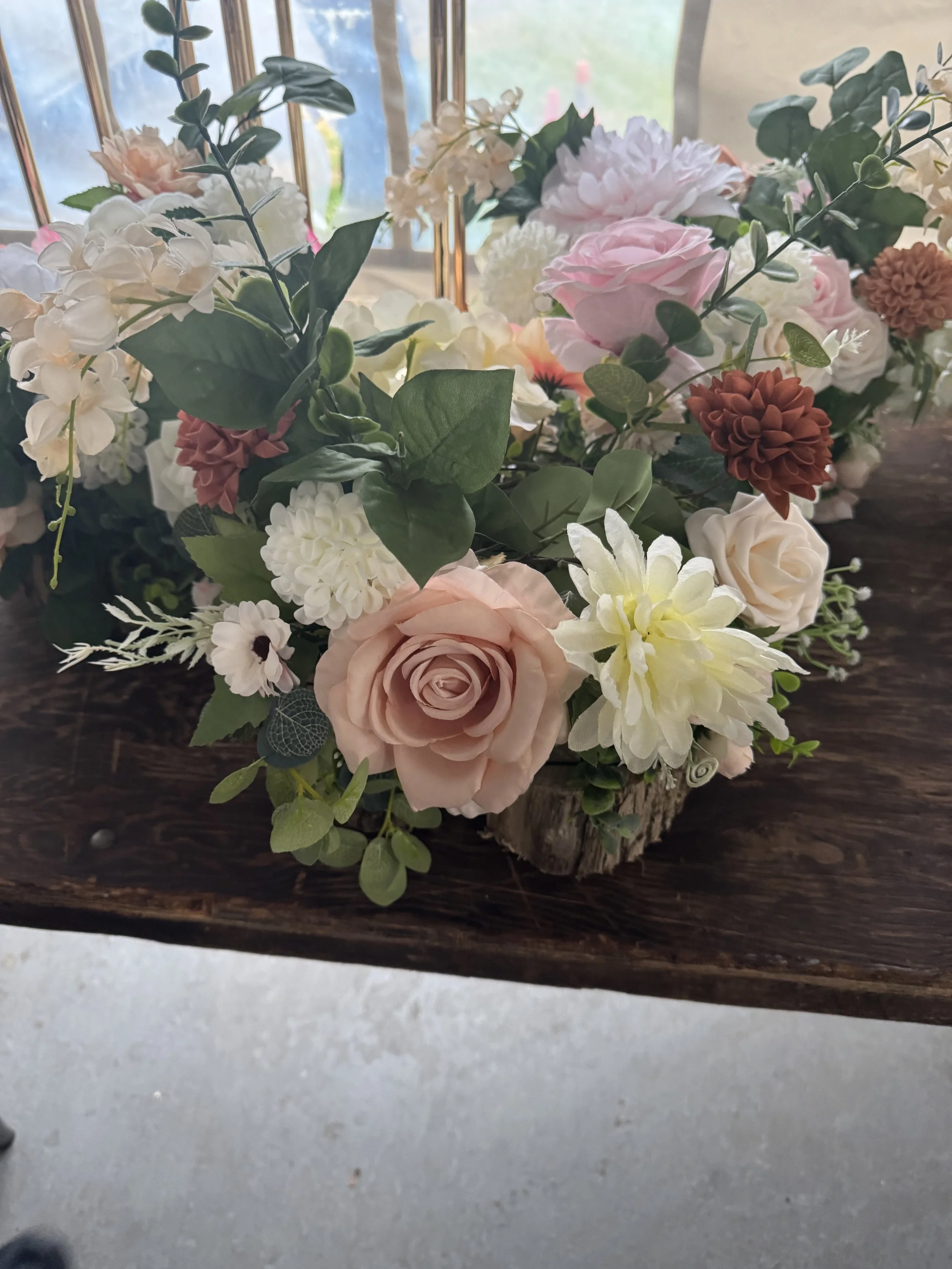 A floral arrangement with pink, white, and brown flowers, including roses and dahlias, placed on a wooden surface near a window.