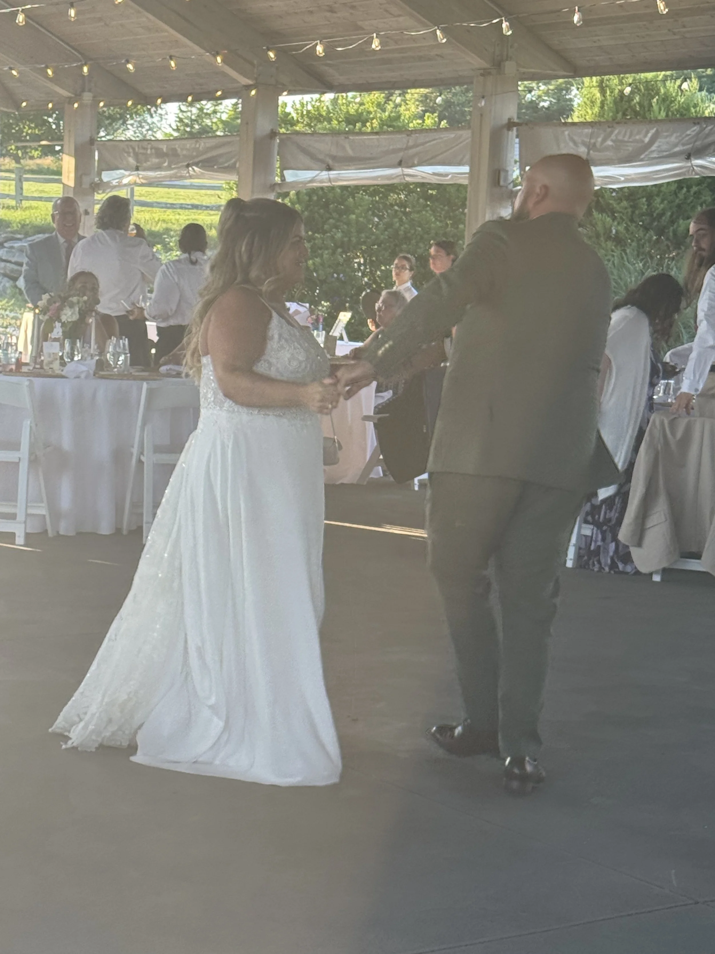 A couple is exchanging vows during their wedding ceremony under a covered outdoor pavilion decorated with string lights, with a long table of guests in the background, and lush greenery visible outside.