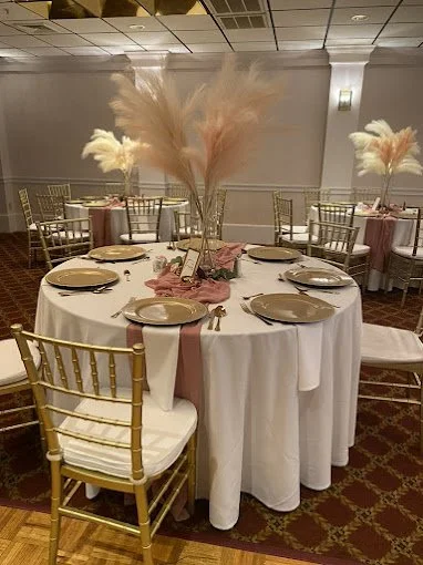 Round banquet table with white tablecloths, gold chairs, pink napkins, and tall decorative centerpieces with feathers.