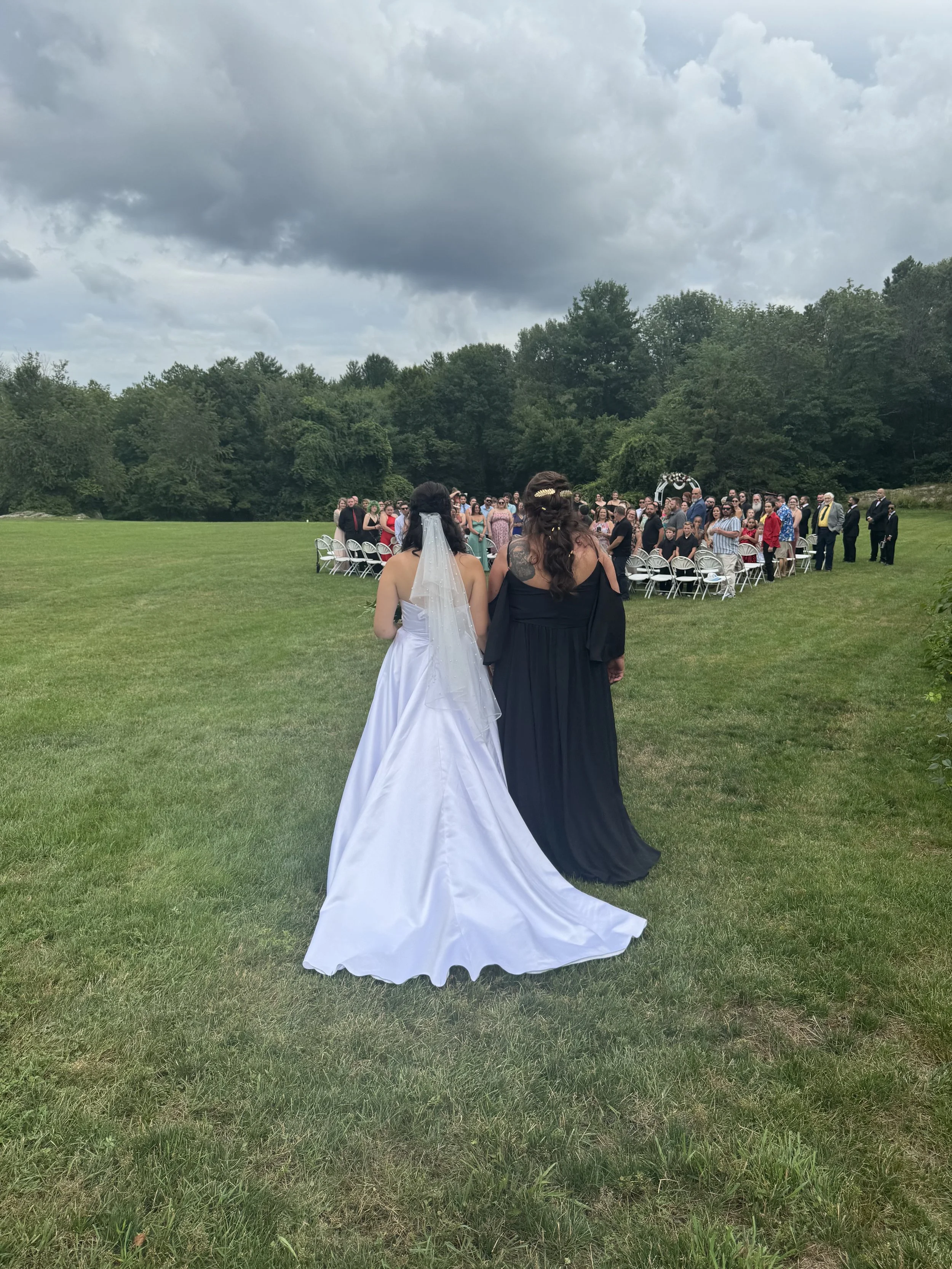 Two women wearing wedding dresses standing on a grassy field, facing a large group of people gathered for a wedding ceremony under a cloudy sky.