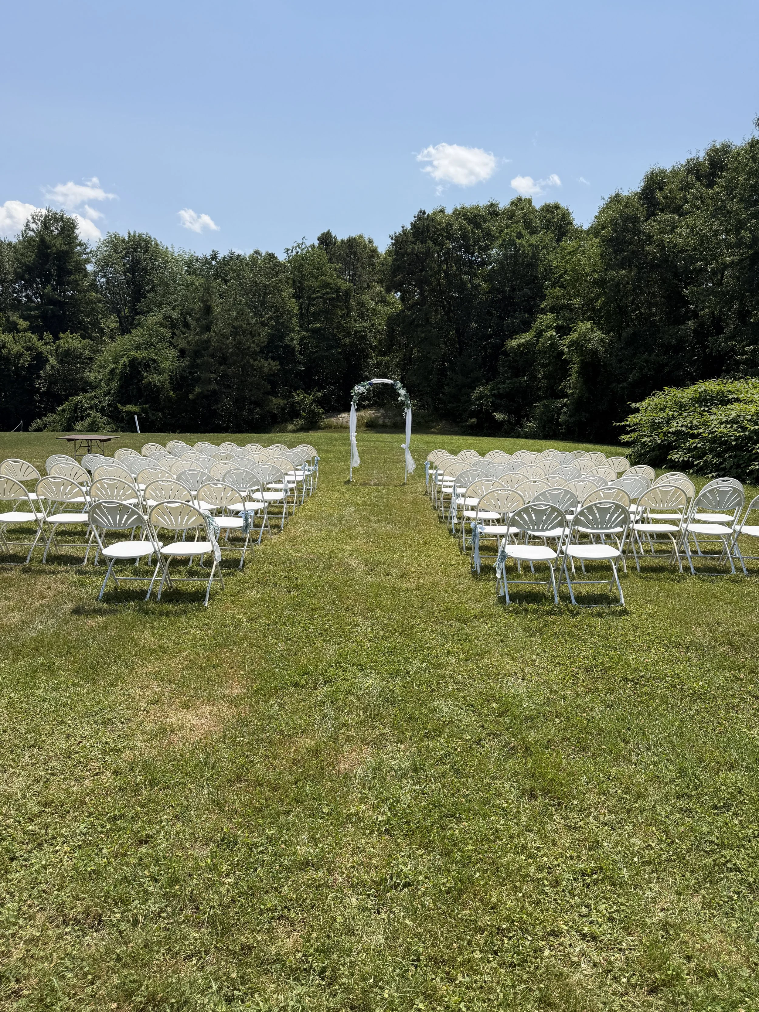 Outdoor wedding ceremony setup with white chairs arranged in rows on a grassy field, facing a white arch decorated with flowers, surrounded by green trees and a blue sky with scattered clouds.