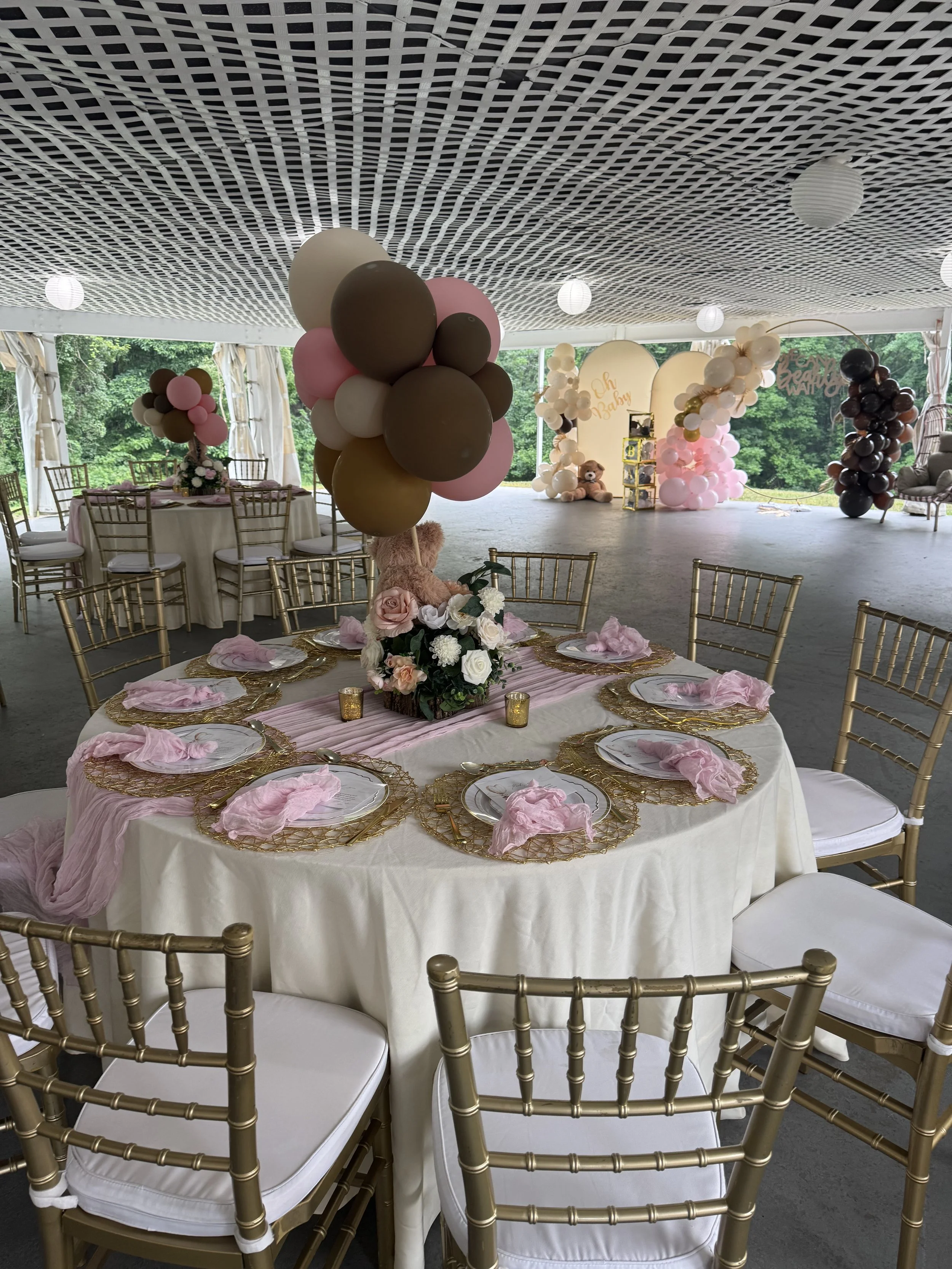 Elegant event space decorated with pink, brown, white, and black balloons, floral centerpieces, and gold accents, featuring round tables with white tablecloths and gold chairs, set up for a celebration such as a baby shower or birthday party.