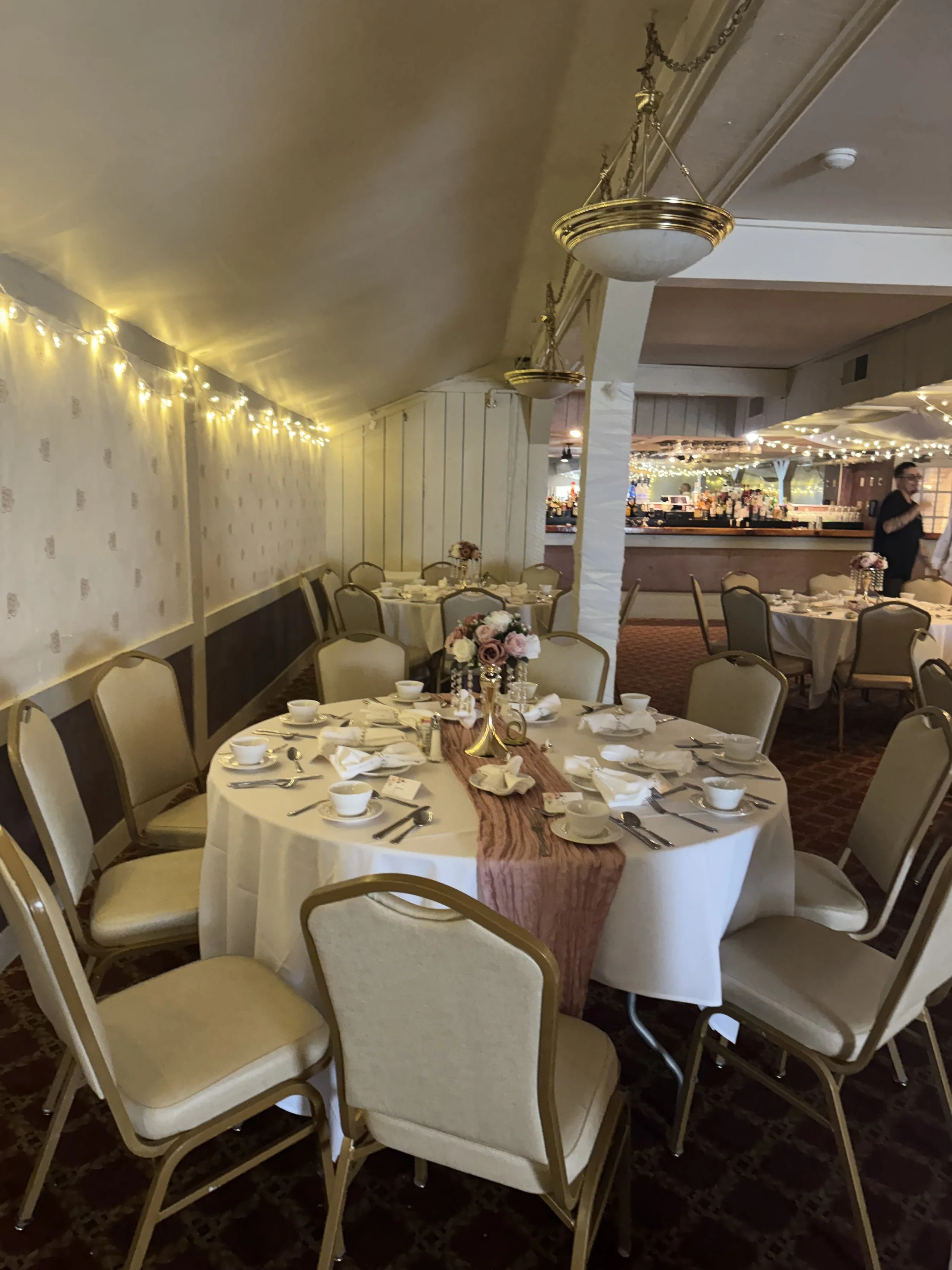 Wedding reception setup with round tables covered in white tablecloths, decorated with pink flower centerpieces, set with plates, cups, and silverware, beige chairs, warm lighting, and a bar area in the background.
