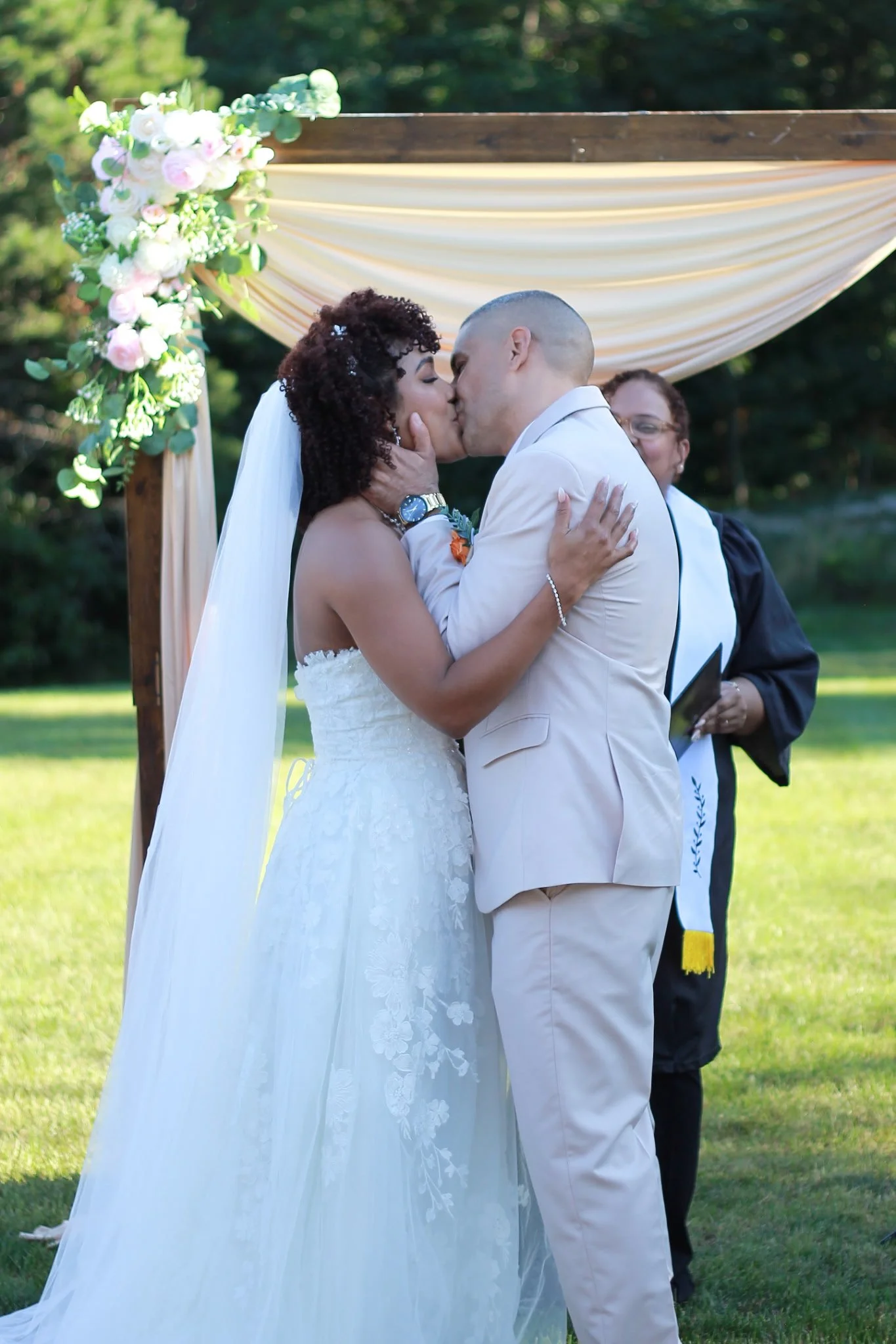 A bride and groom share a kiss during their outdoor wedding ceremony under a decorated arch with flowers and drapes, with an officiant in the background.