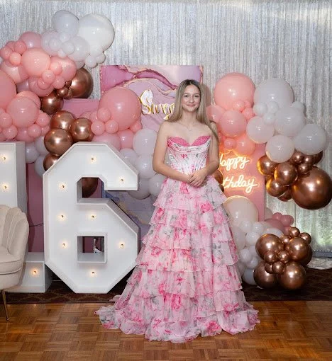 Young woman in pink and white floral dress standing in front of birthday decorations with pink, white, and bronze balloons, a large illuminated number 6, and a neon 'Happy Birthday' sign.
