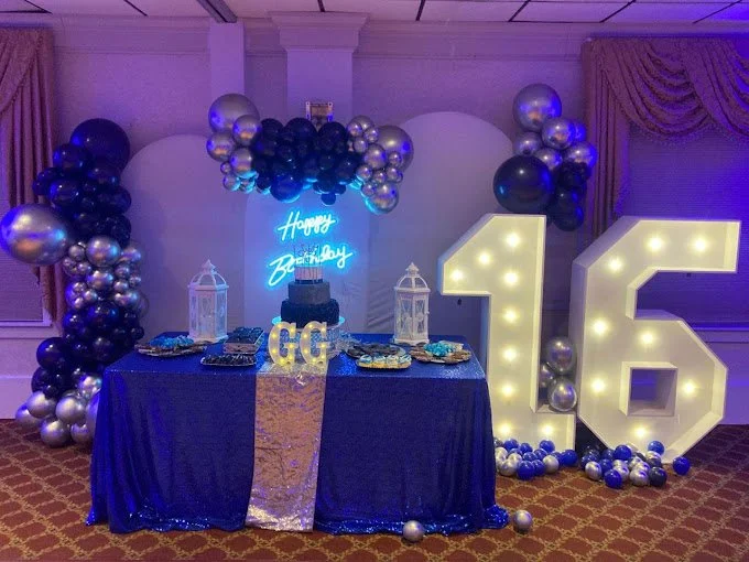 Birthday party setup with a blue and silver theme, featuring a decorated table with snacks, a cake, and a neon sign that says 'Happy Birthday.' There are large illuminated number '16' and a balloon arch overhead.