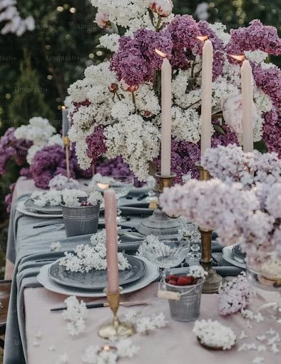 Elegant outdoor table setting with pink and white flowers, tall pink candles, and silver dishes and glasses for a formal event.
