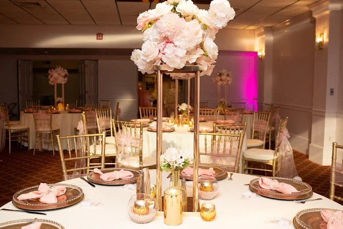 Elegant wedding reception table with gold chairs, pink napkins, floral centerpieces, and soft lighting in the background.
