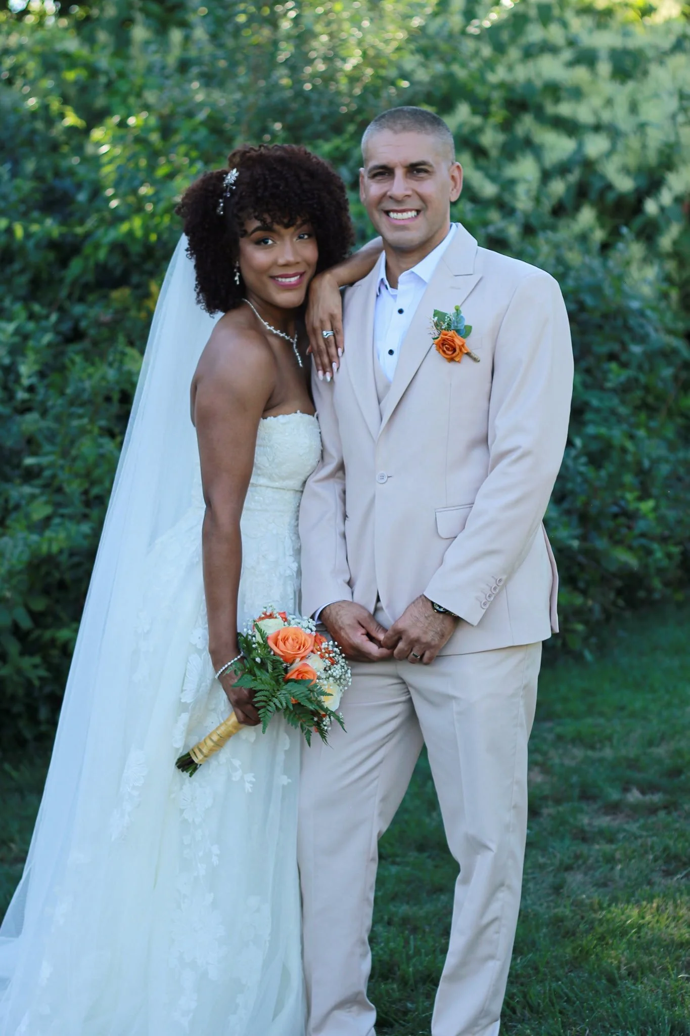 A newlywed couple standing outdoors in a green, leafy area. The bride, with dark curly hair, is wearing a white strapless wedding dress and earrings, holding a bouquet with orange roses. The groom, with short gray hair, is dressed in a beige suit wit