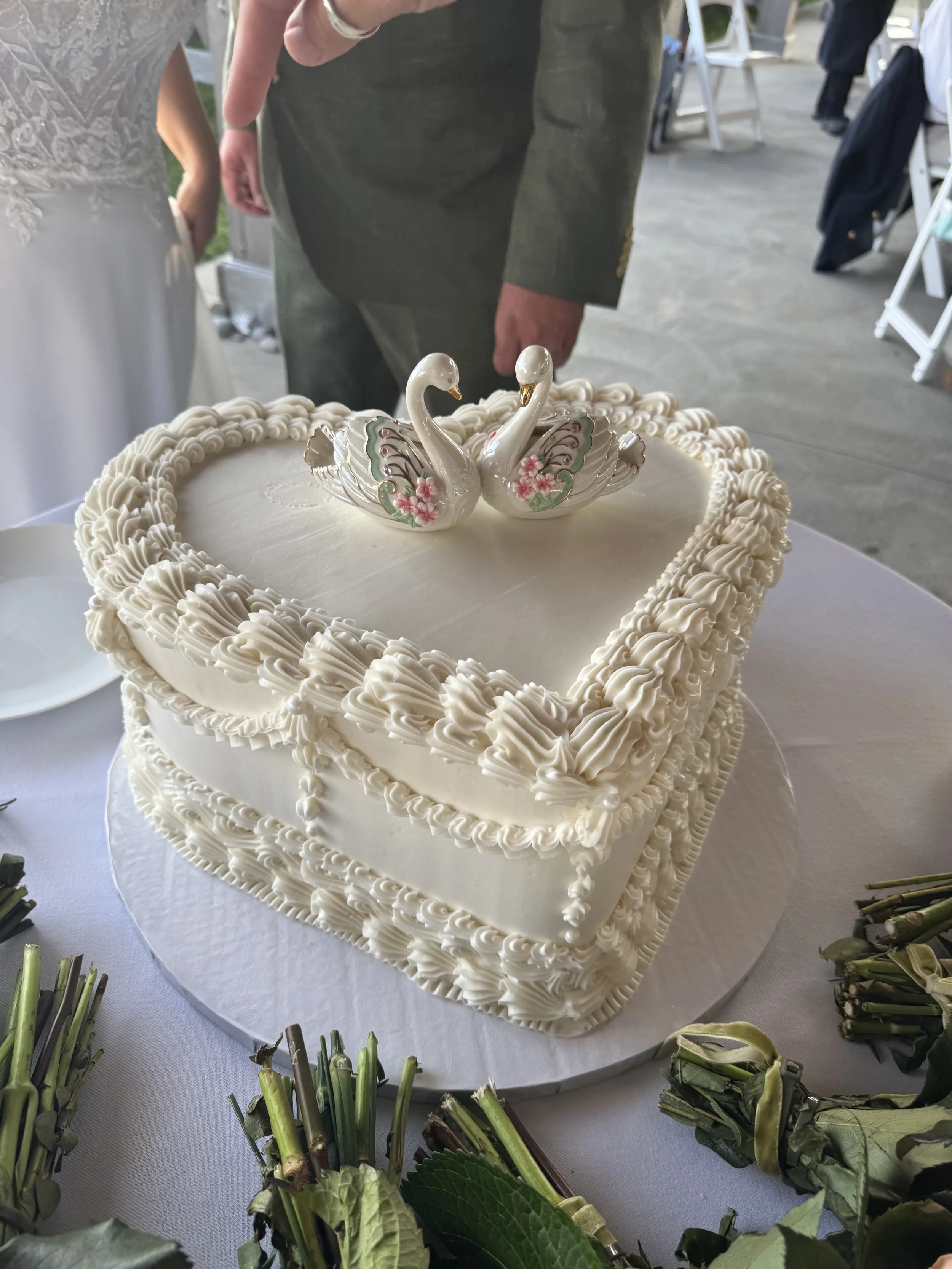 A wedding cake with two swan decorations on top, surrounded by green floral arrangements. The cake has white icing with piped borders. People in formal attire are present in the background.
