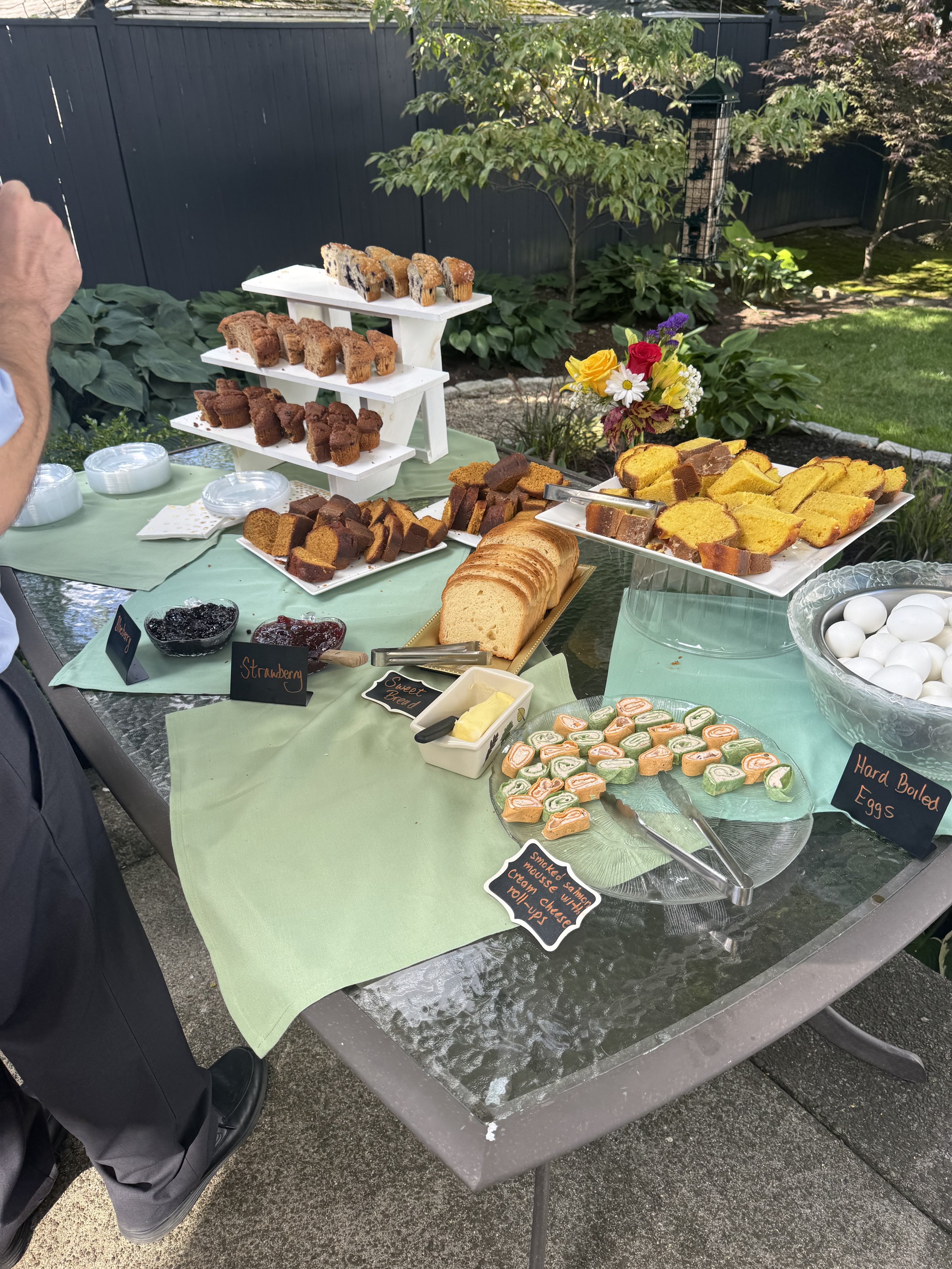 A dessert table outdoors with various baked goods, including slices of bread and cakes, some labeled and displayed on plates and stands. There are signs with handwritten labels for treats such as strawberry, sweet bread, smoked salmon mousse with che