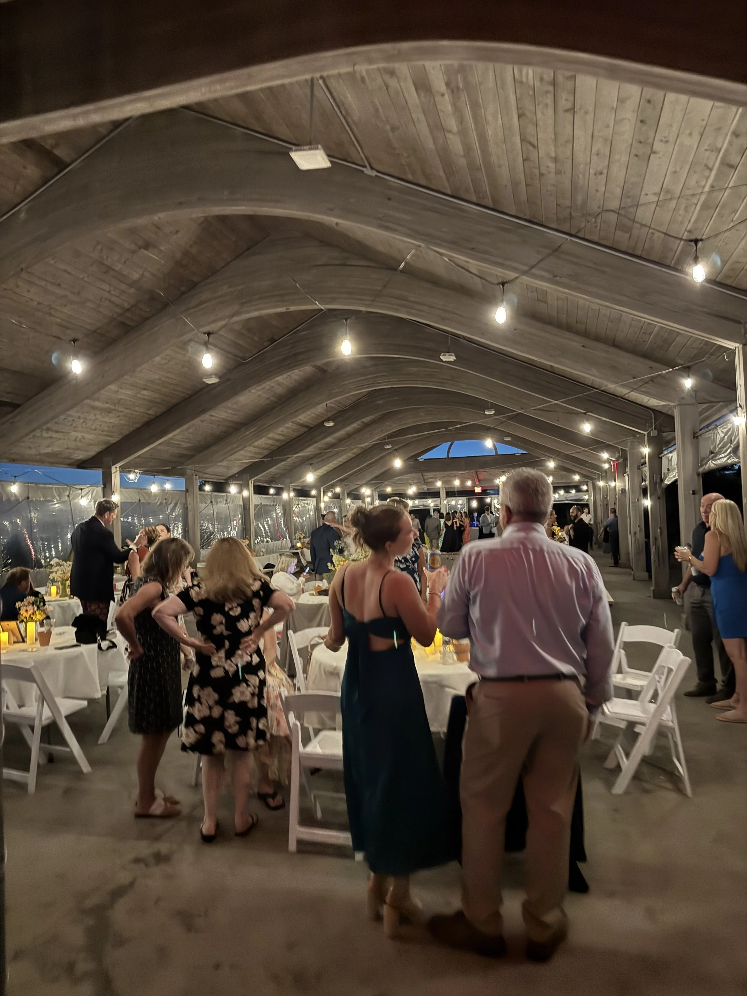 People socializing at a formal event in a large, covered outdoor space with string lights and tables