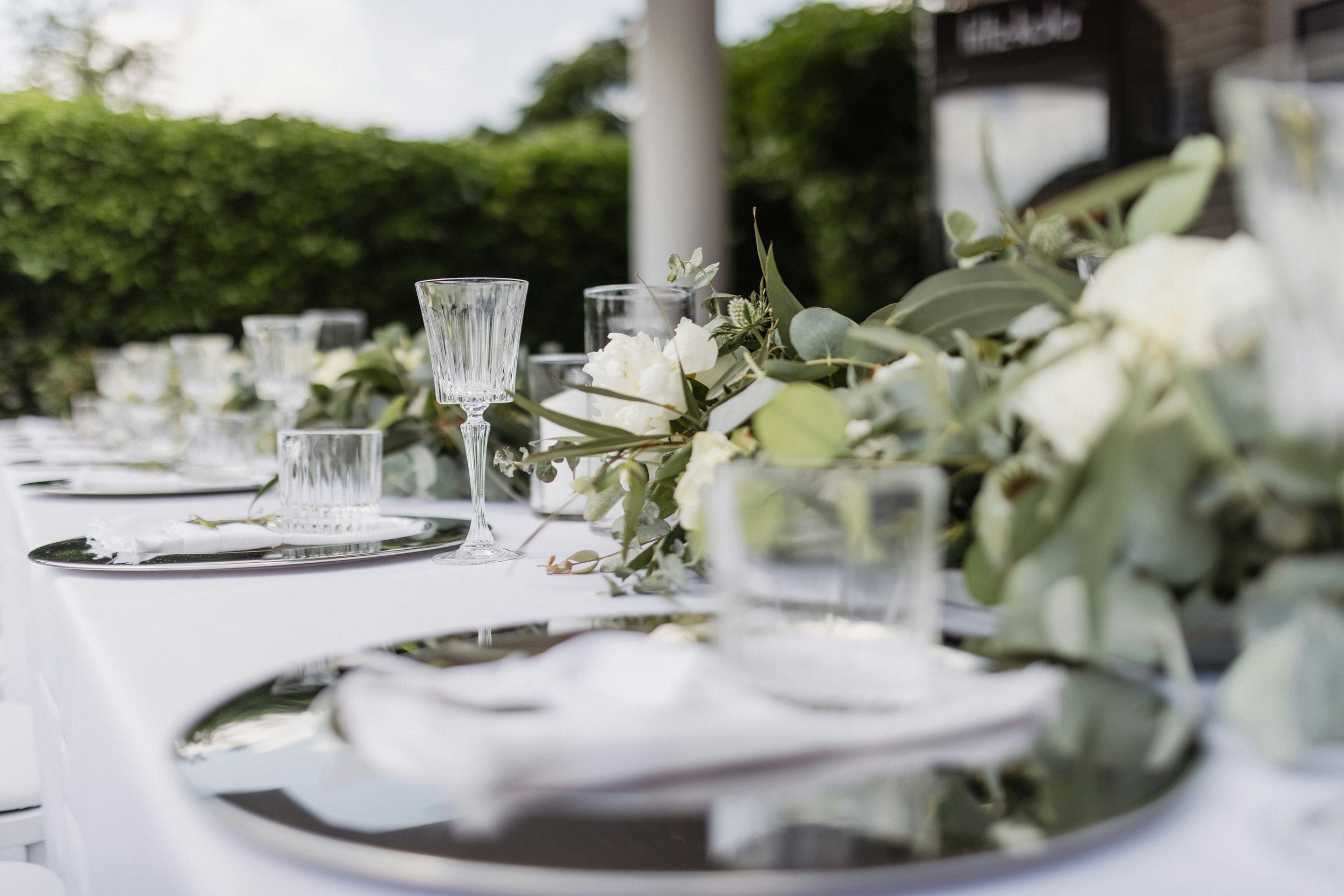 A table set outdoors with glassware and floral arrangements, including white flowers and green leaves, against a background of greenery.