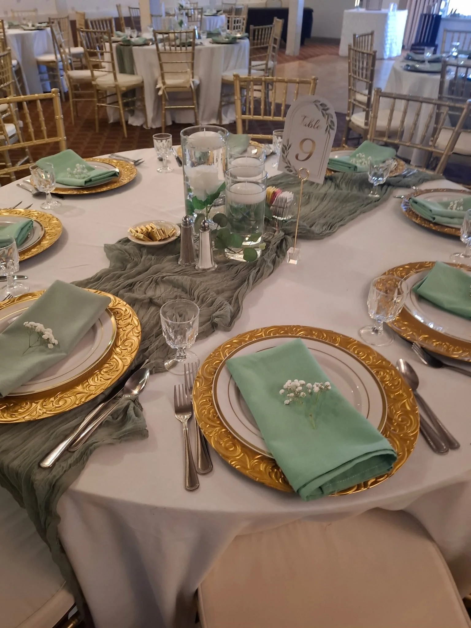 Wedding reception table decorated with gold-rimmed chargers, mint green napkins, and centerpieces with candles and greenery.