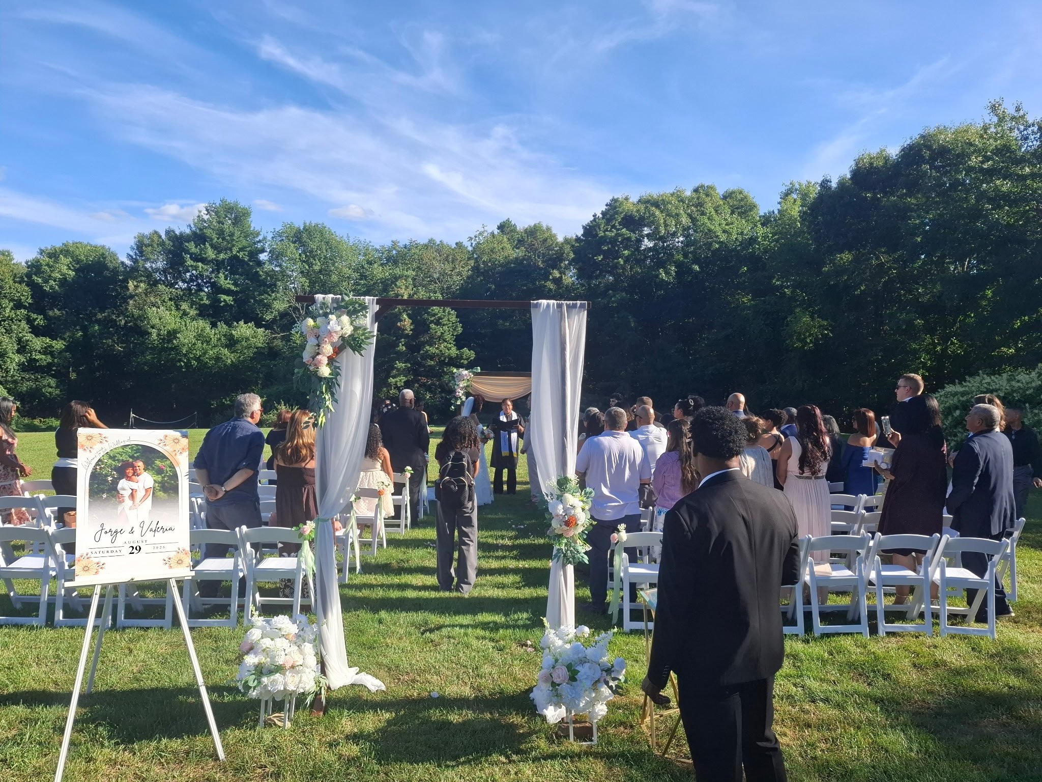 An outdoor wedding ceremony with rows of white chairs decorated with floral arrangements, a wooden arch with white drapes and flowers, and guests standing on a grassy field under a blue sky with some clouds, surrounded by trees.